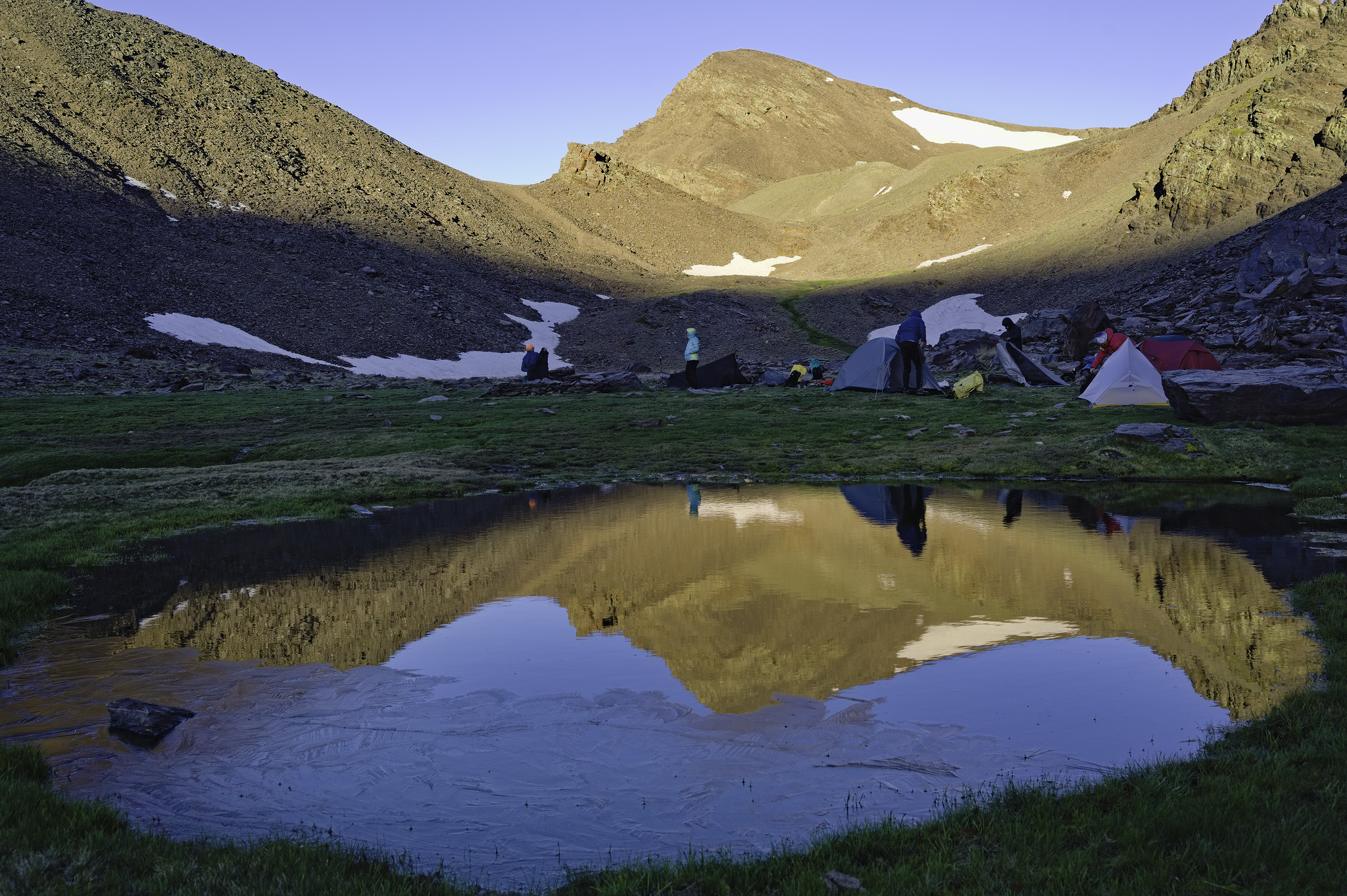 A mountain peak is illuminated by morning sunlight, which is reflected perfectly in a small round lake surrounded by grass. In the middle are a few small tents