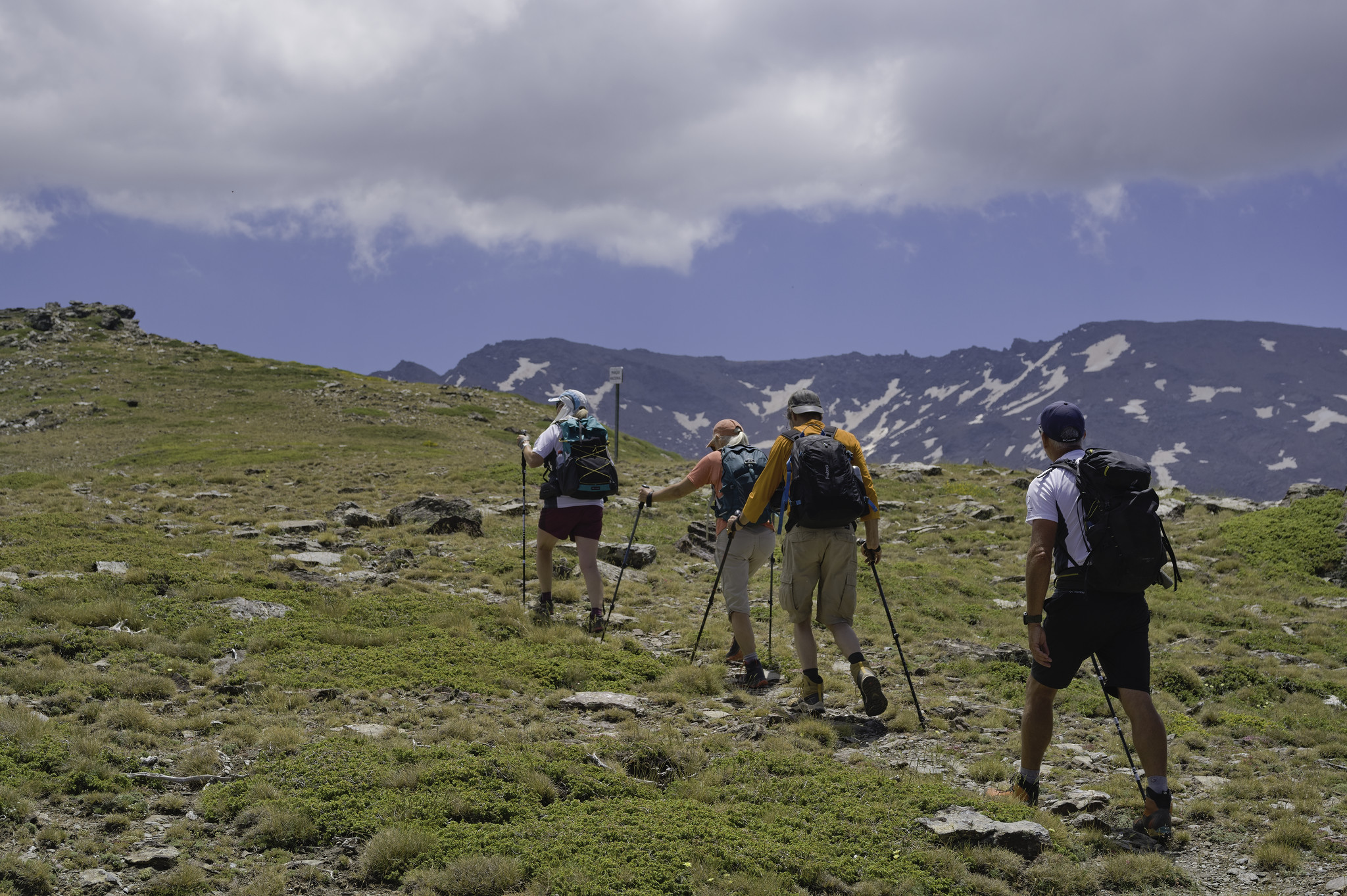 4 hikers pass along a broad ridge with a mountain range with some snow patches to the right