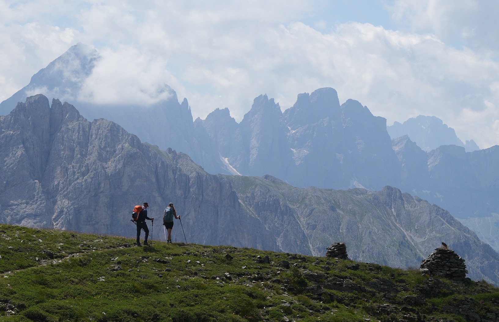 Two hikers walk along a trail with jagged mountain scenery behind