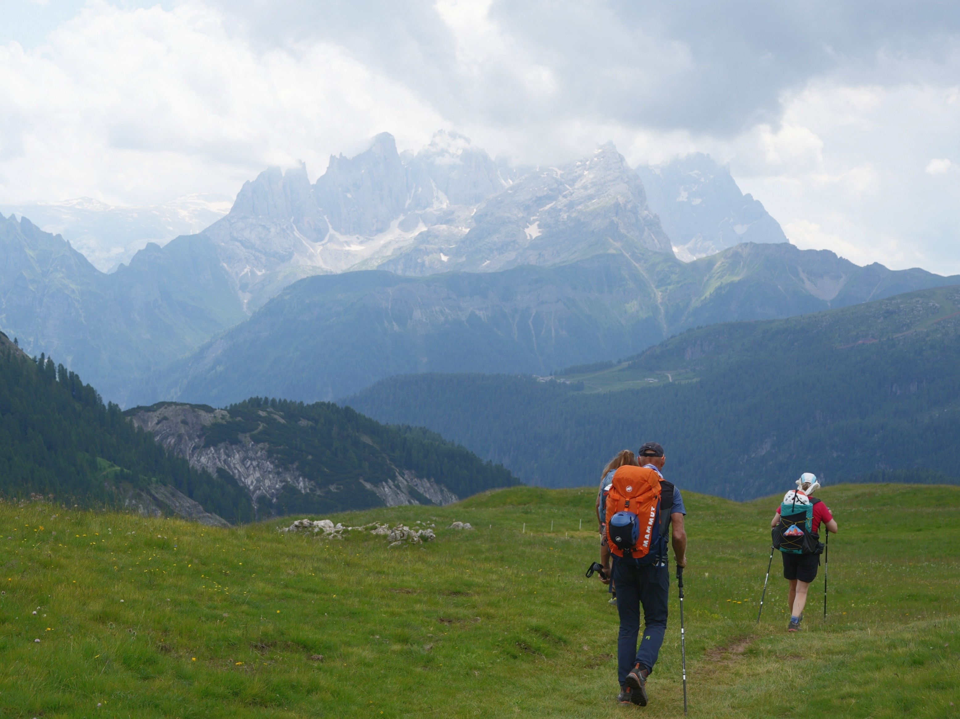 Twp hikers walk through a green meadow with some jagged mountains behind them