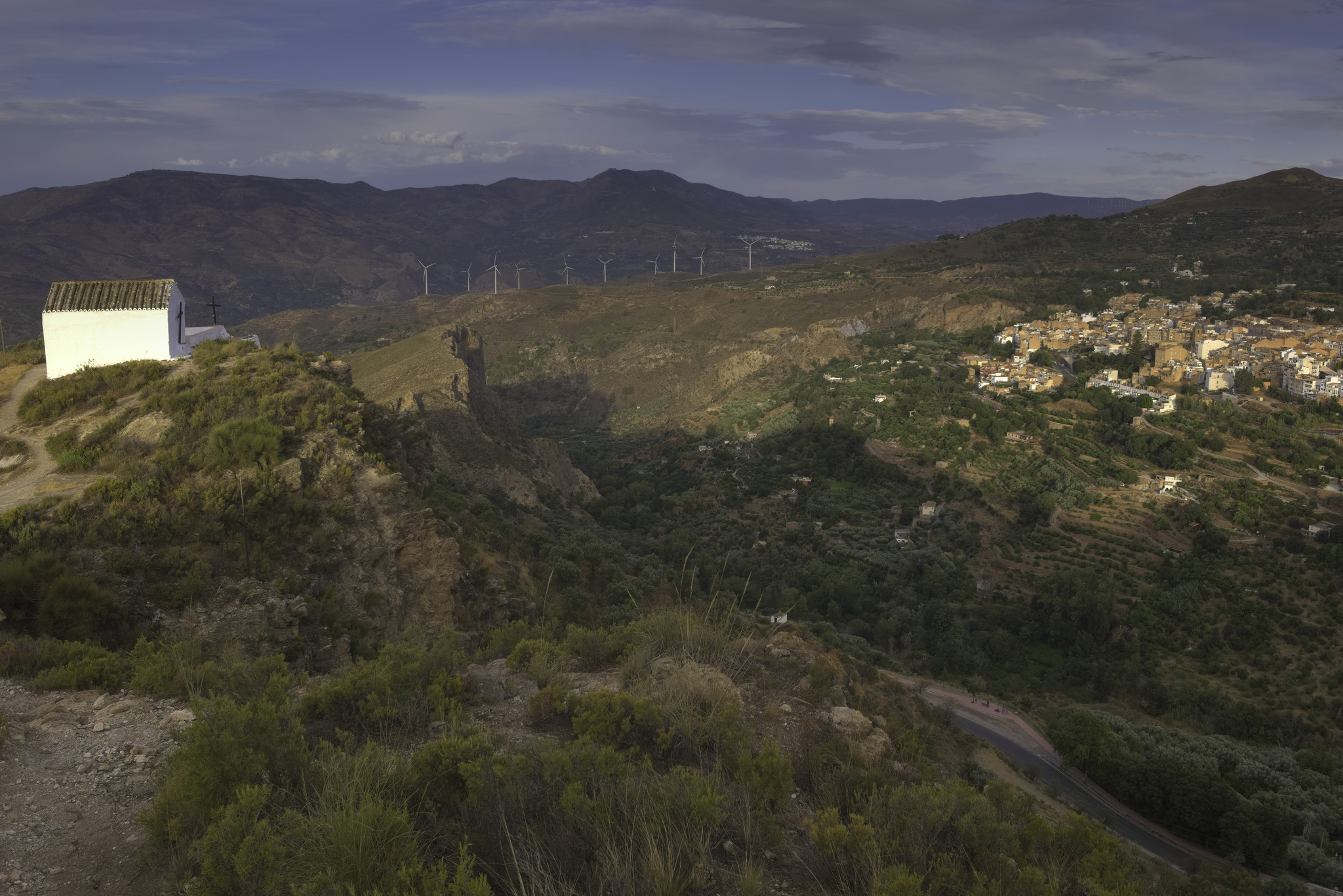 A white chapel on a hill on the left overlooks a green valley with a small town sitting on the right hand side on a plateau