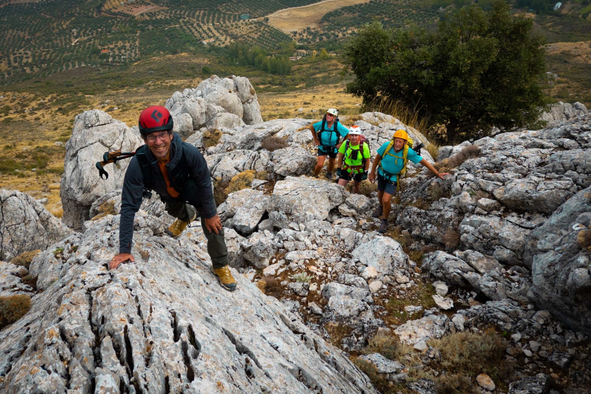 A person with red helmet looks up at the camera whilst others make their way up to him. They are climbing a rocky hillside