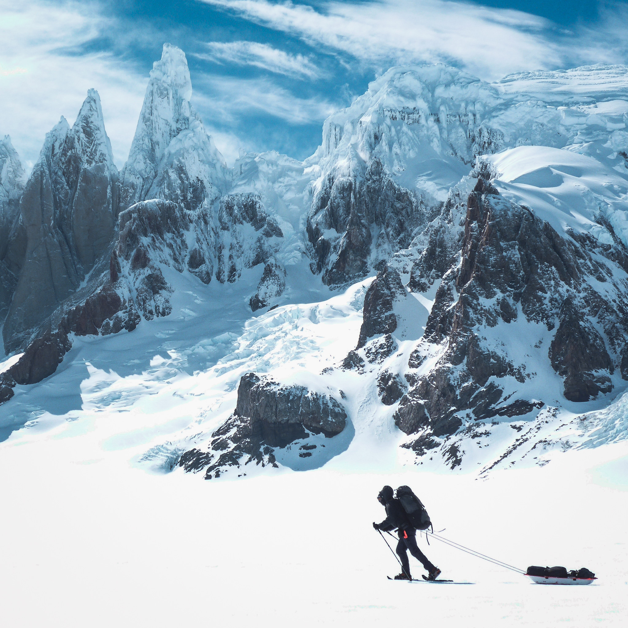 A lone figure pulls a sledge across a snowy glacier. Behind him rise some jagged mountains covered in ice