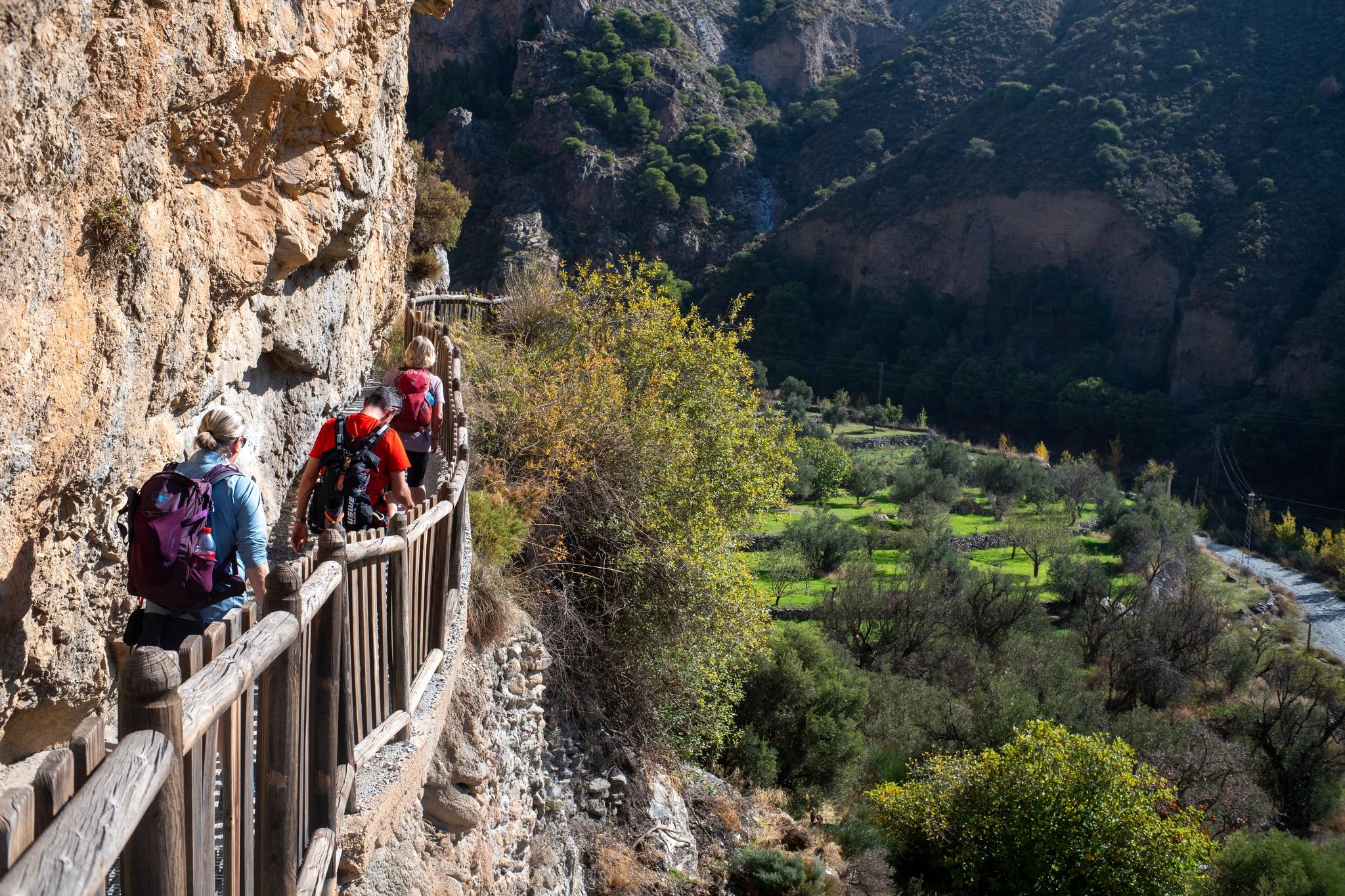 Left hand side is a narrow walkway on the Ruta de Pavilla with three hikers walking along it. A green valley to the right