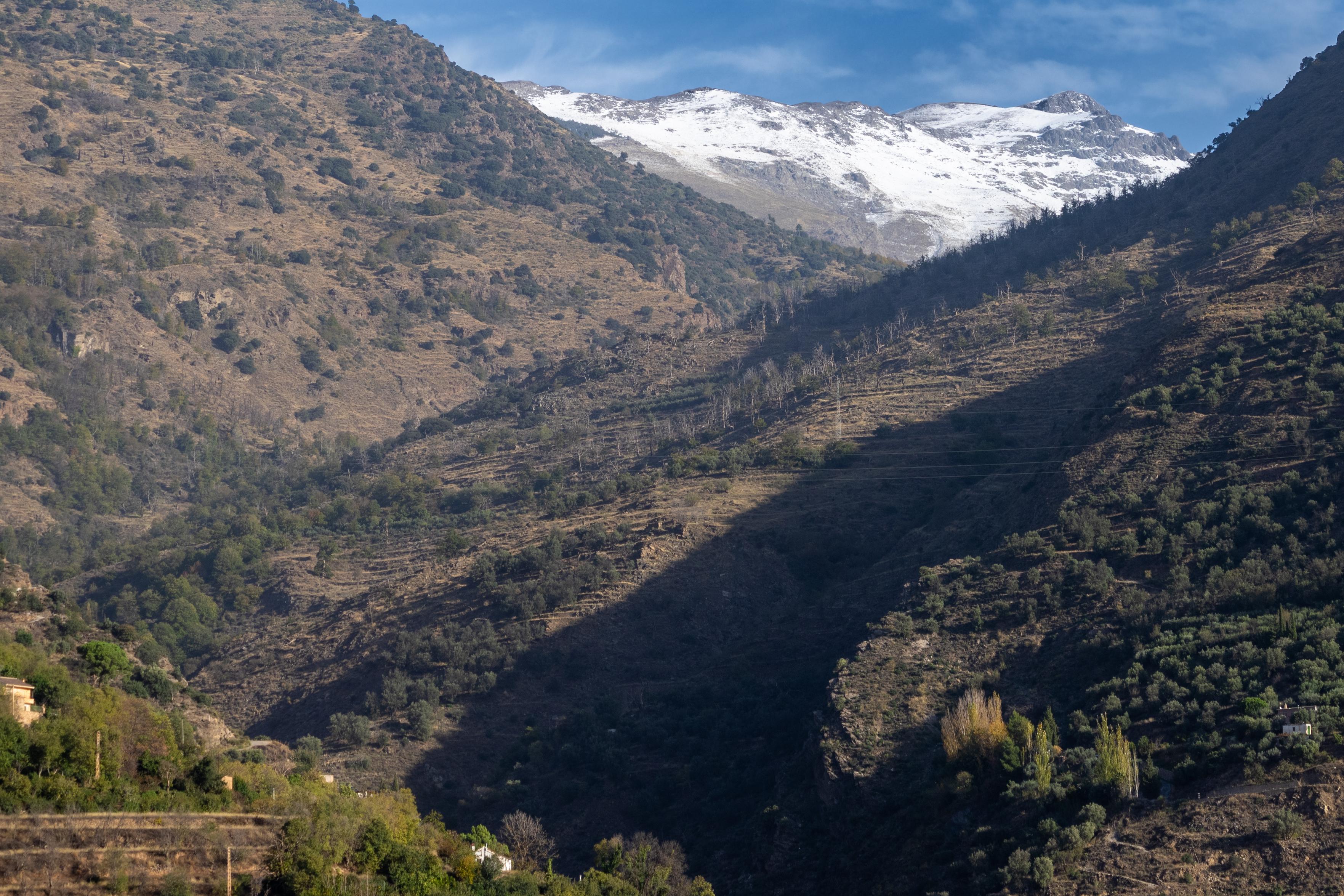 A valley winds it's way upwards through lush green and brown hillsides until it meets snow on a high mountain summit