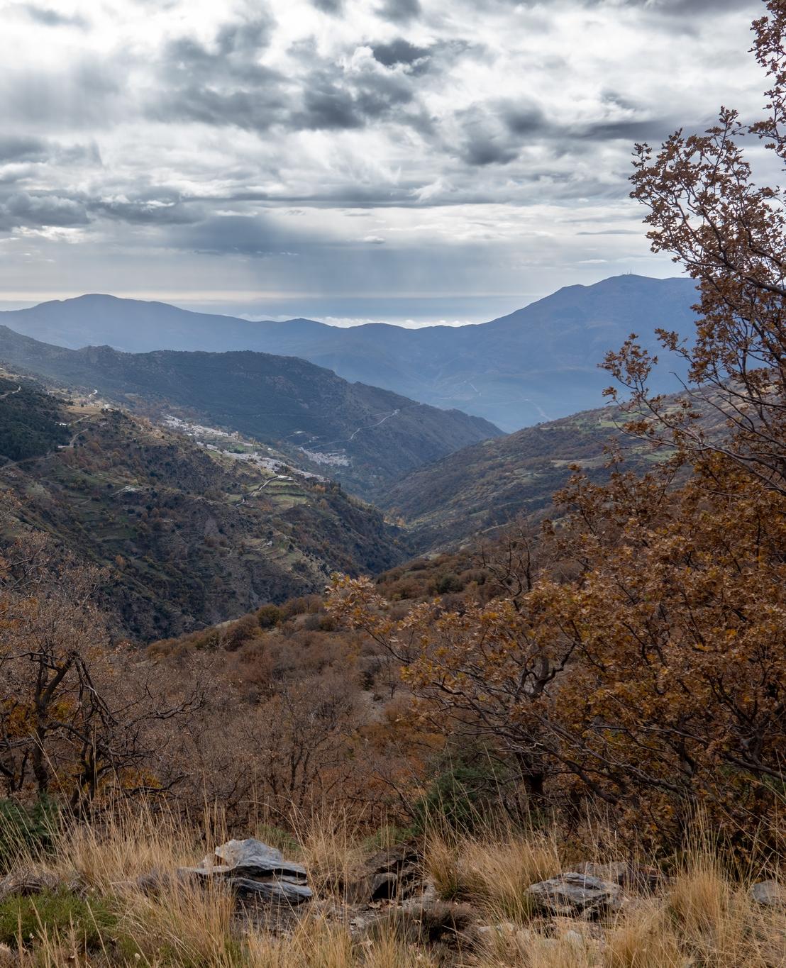Looking back down the Poqueira Valley to the town of Capileira perched on the hillside. Rays of sun and coming down through the dark clouds. Autumn colors on the trees and shrubs in the foreground