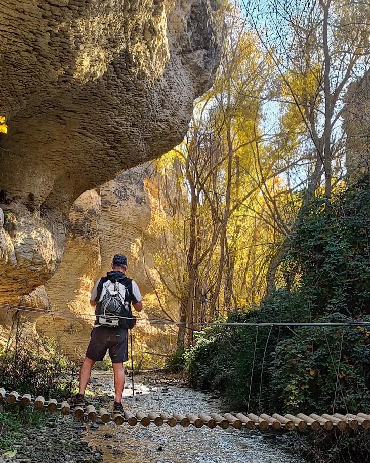 Person on a rikkety bridge over a small river with huge rock walls towering above and a very colorful scene upstream.