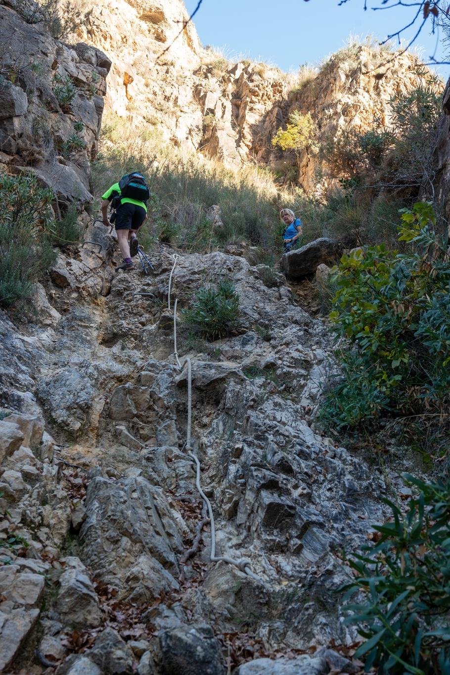 A person in yellow starts to descend a rocky section of path alongside an old rope