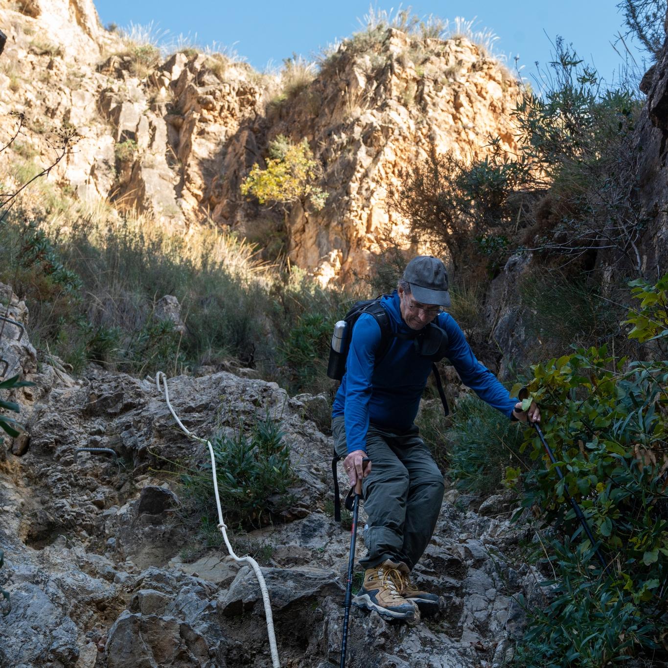 A person in blue starts to descend a rocky section of path alongside an old rope