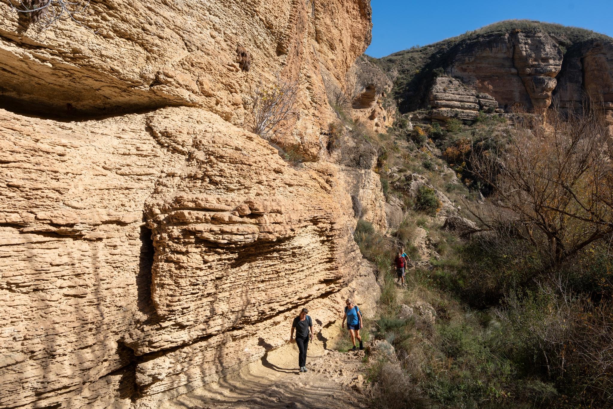 some hikers pass by the base of a huge yellow cliff to the left