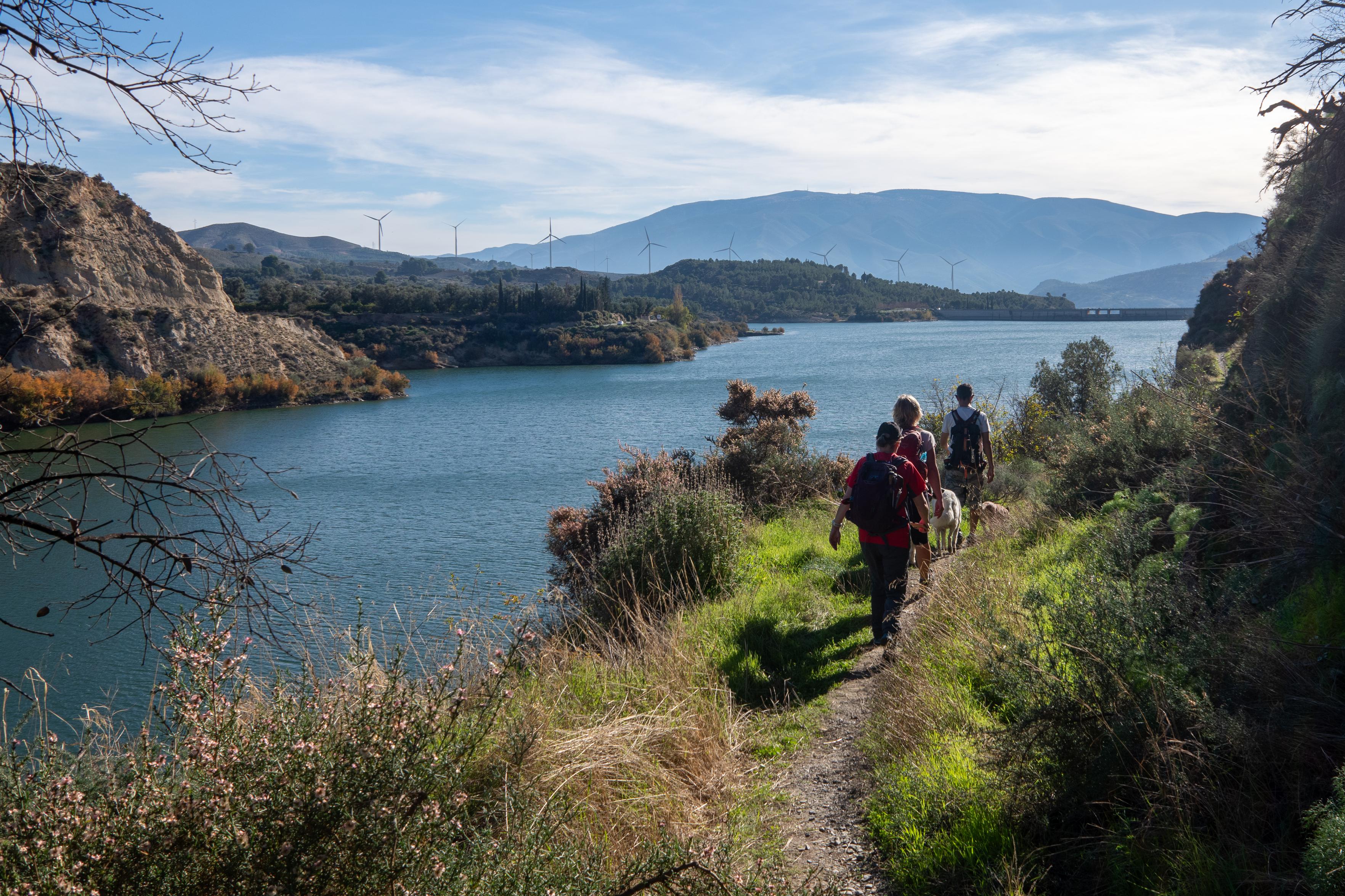 People walk along a narrow trail with a lake to the left
