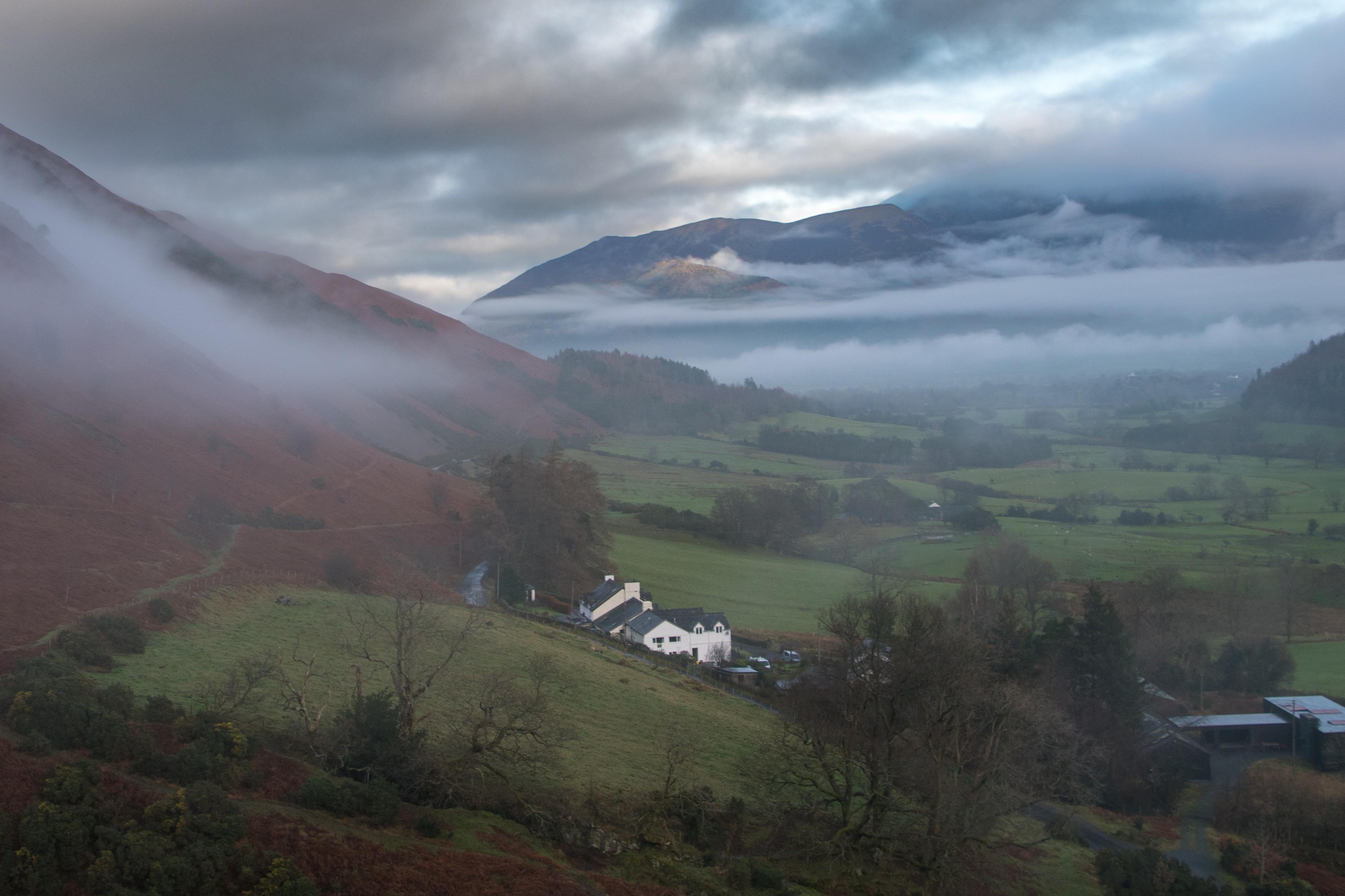 Looking down the Newlands Valley to the peak of Skiddaw which is enclosed in cloud. A famhouse in the centre has red heather and bracken slopes going up to the left. Low inversion mists cloud the hillsides