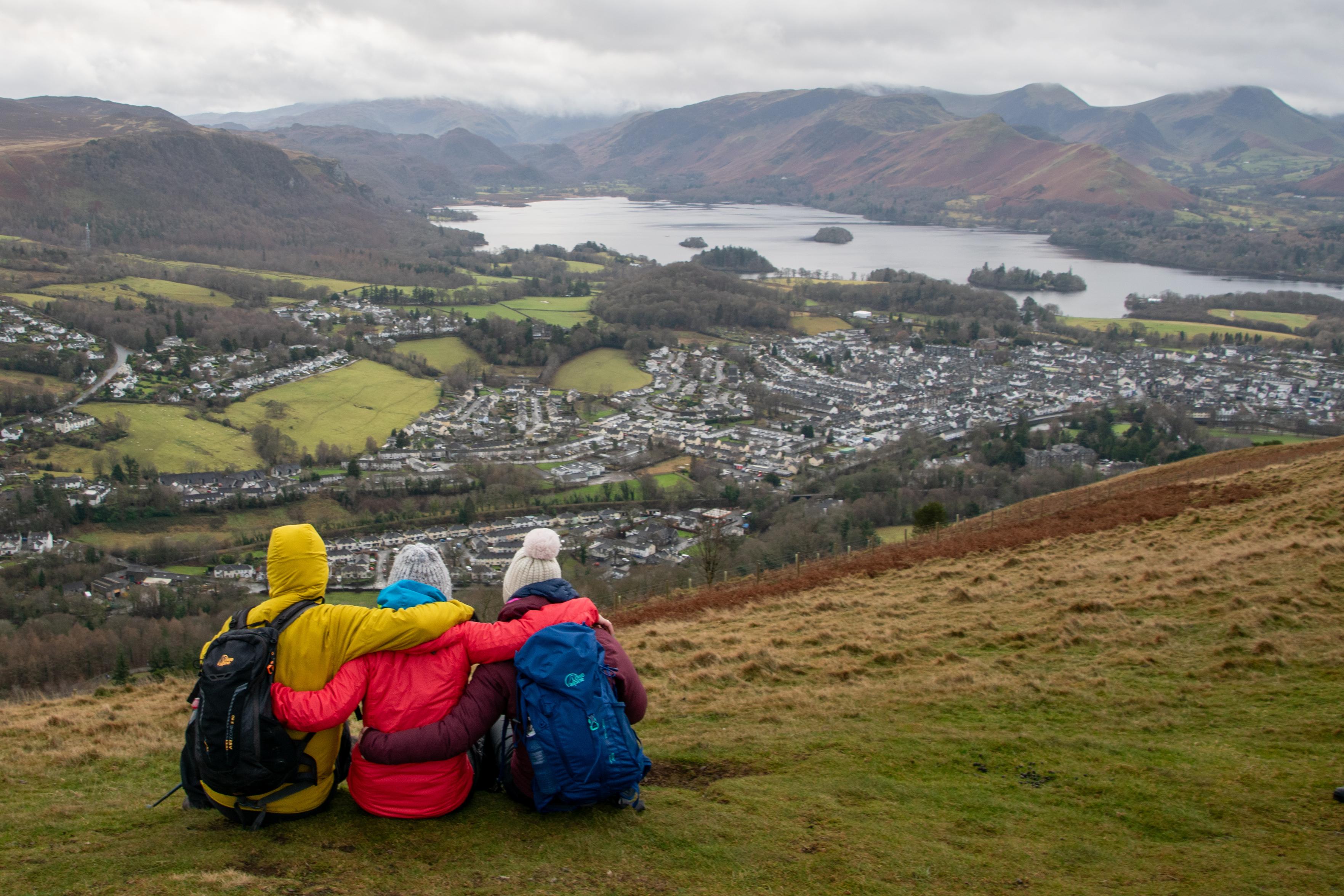 Three people in multi-colored jackets sit with their arms round each other looking over a town lake and hills.