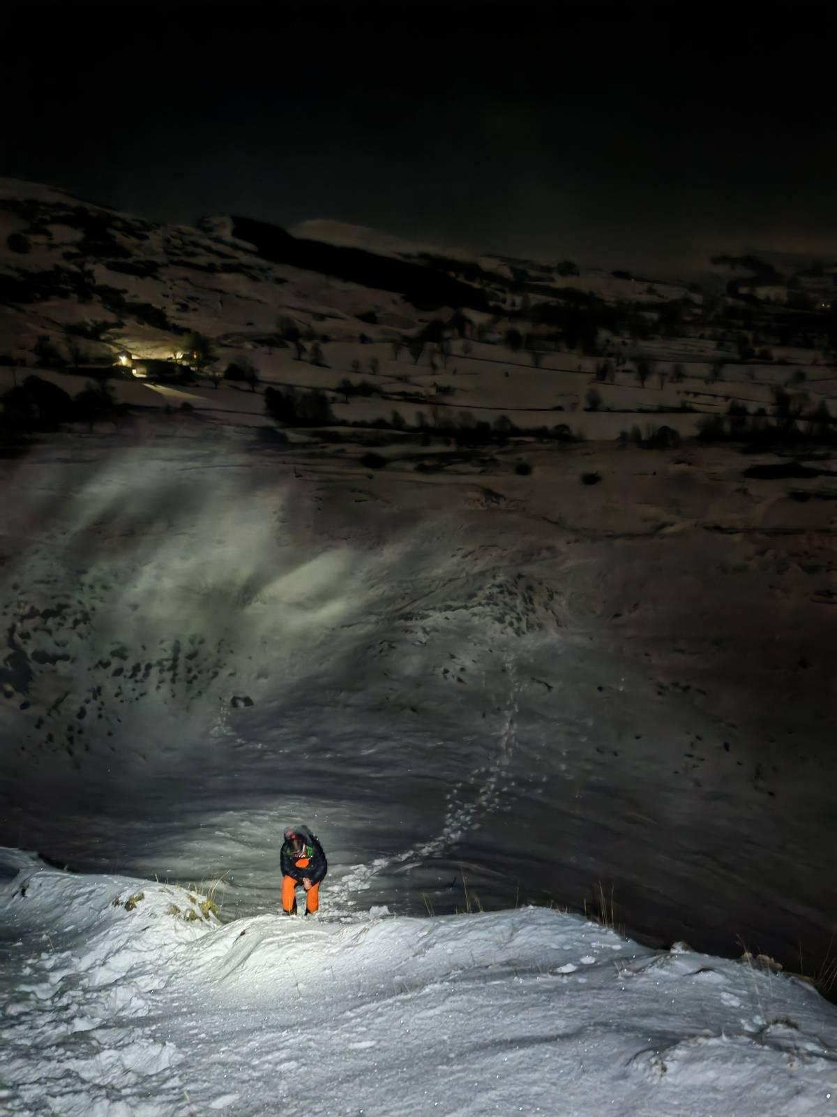 A person in orange pants is in the snow in the foreground illuminated by head torch. Behind is a dark snowy scene with only some lights from a farmhouse showing