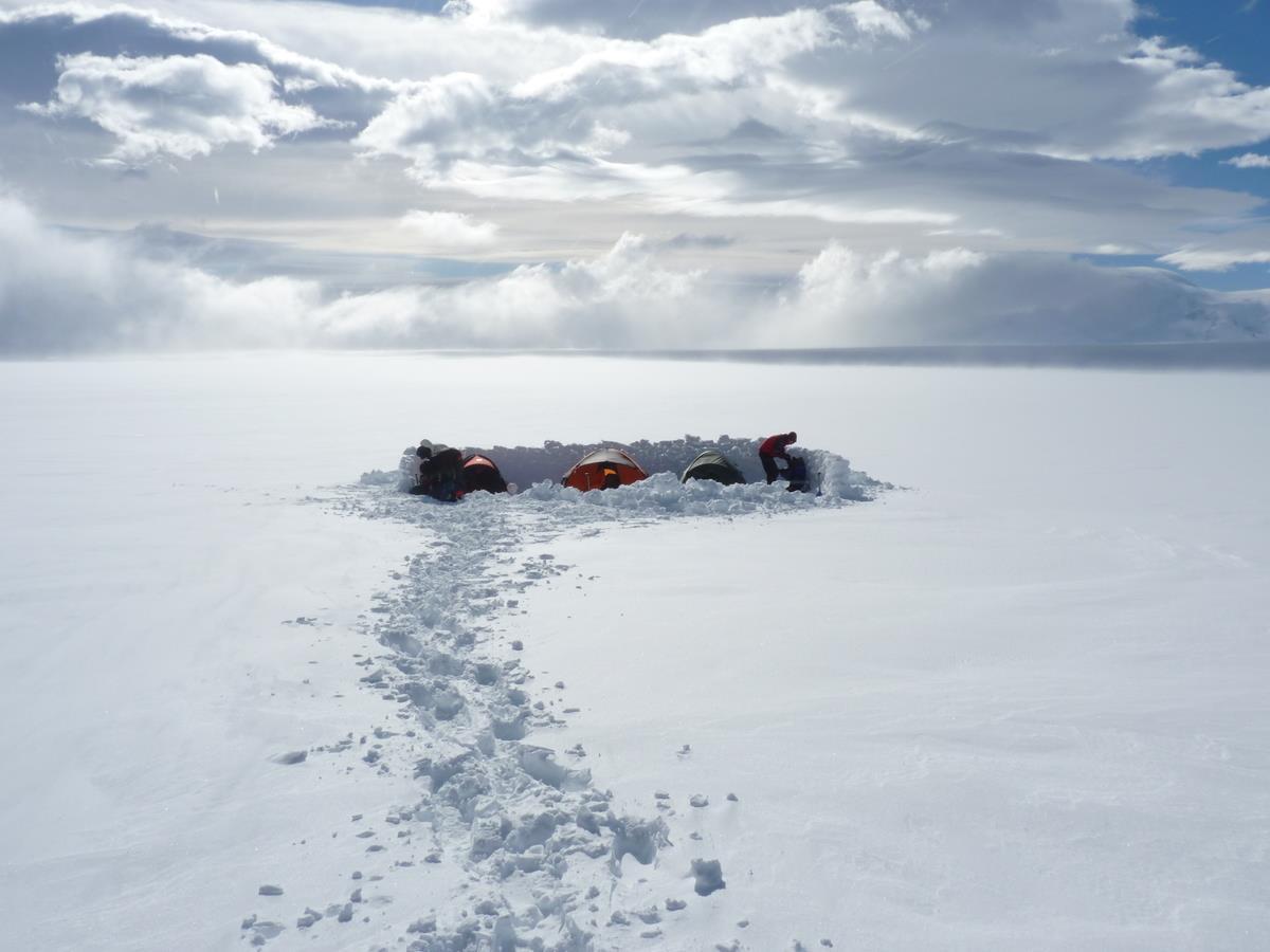 A remote camp site surrounded by snow and stretching into the distance. Around a small number of tents are built snow walls to resist the string patagonian winds.