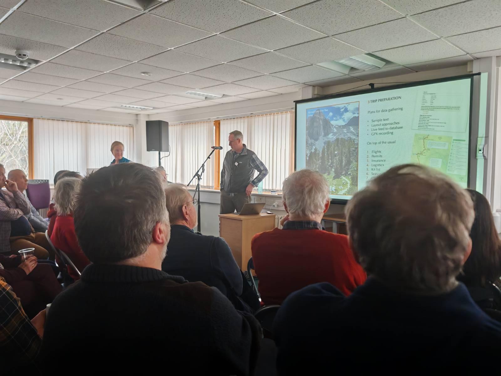 A group of people listen to a speaker with a large screen display to the right