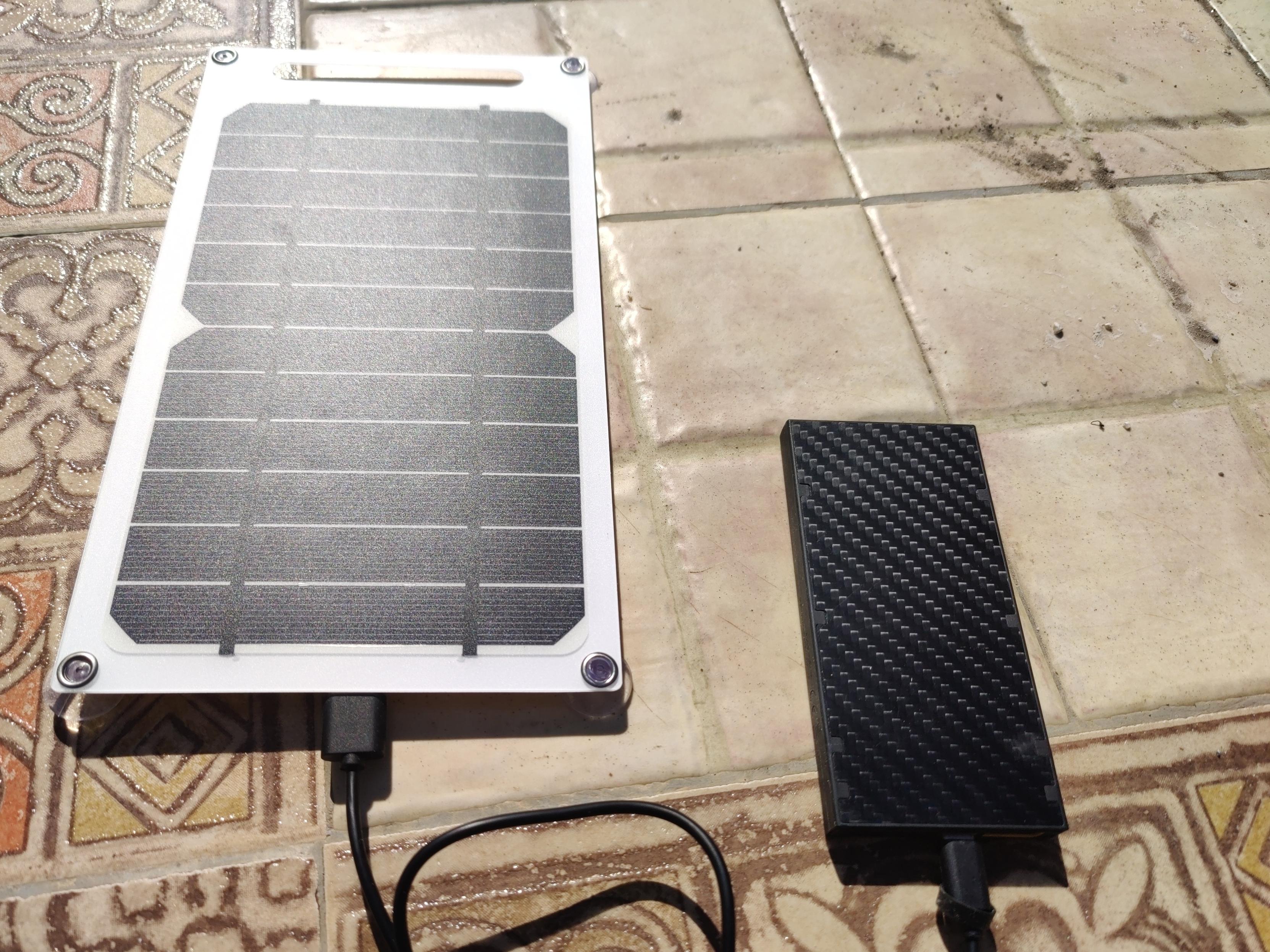 A solar panel on a table charges up a small power bank using a black cable