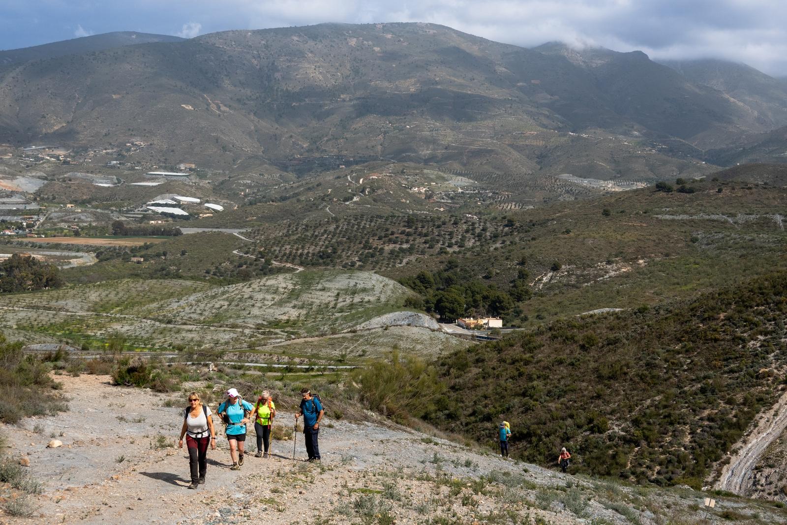 4 hikers climb a broad path leading up a mountain with some rolling hills behind