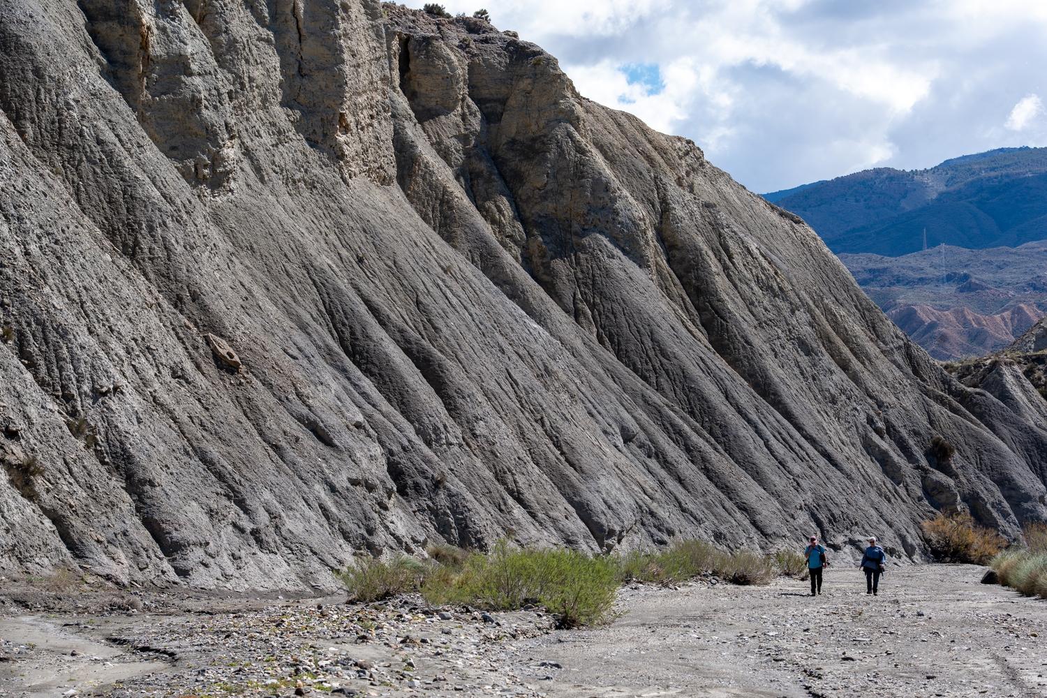 A huge loose wall of crumbling stone rears above two hikers on the path below and to the right