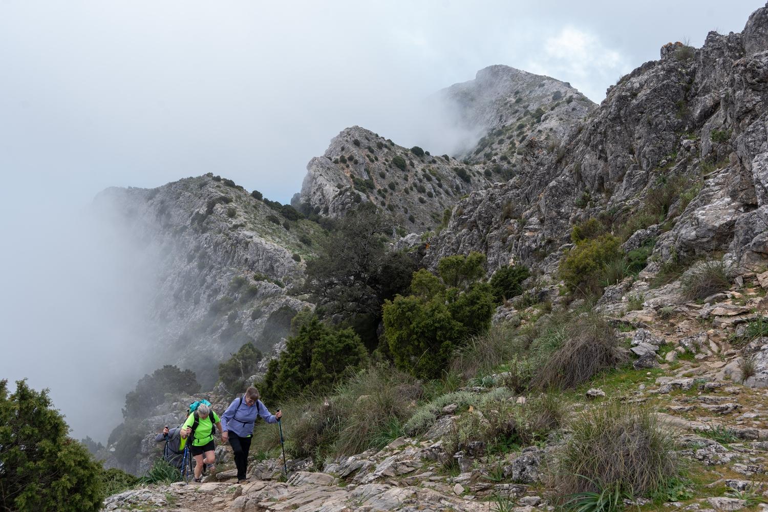 An undulating ridge with mist and clouds to the left. In the foreground to the left are some hikers ascending