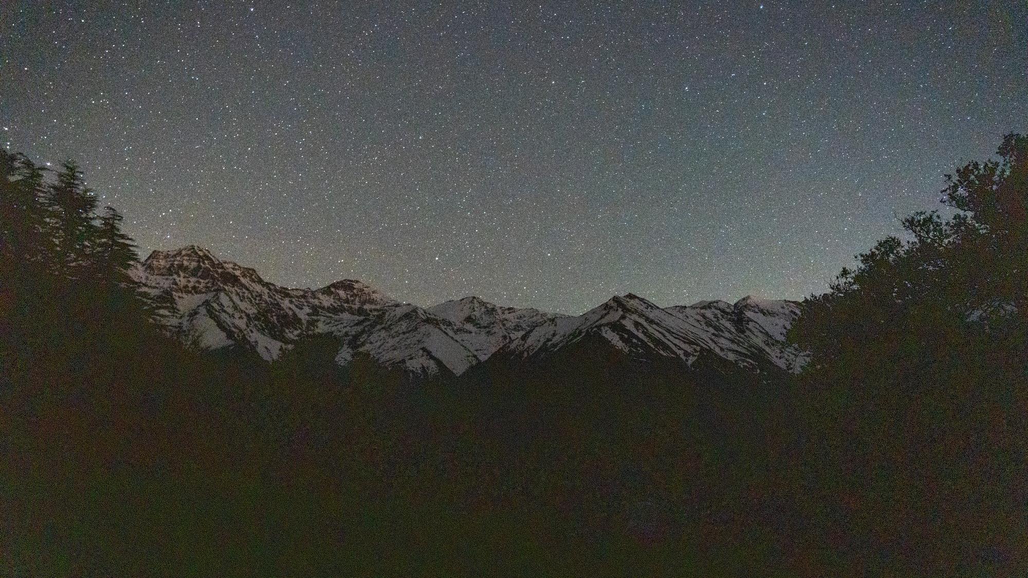 A grainy night image of some snowy mountains with hundreds of stars above, the sides lined by trees