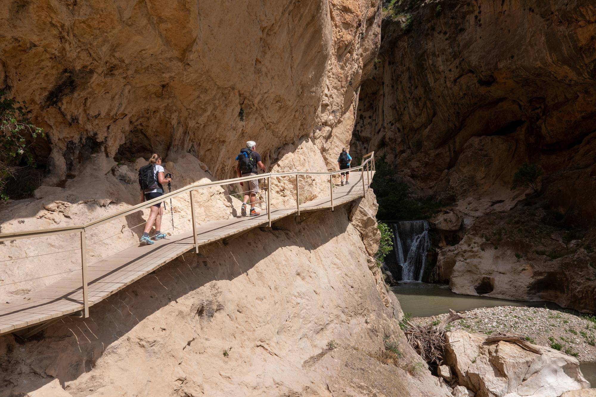 3 hikers walk along a wooden elevated platform alongside cliffs and above a river