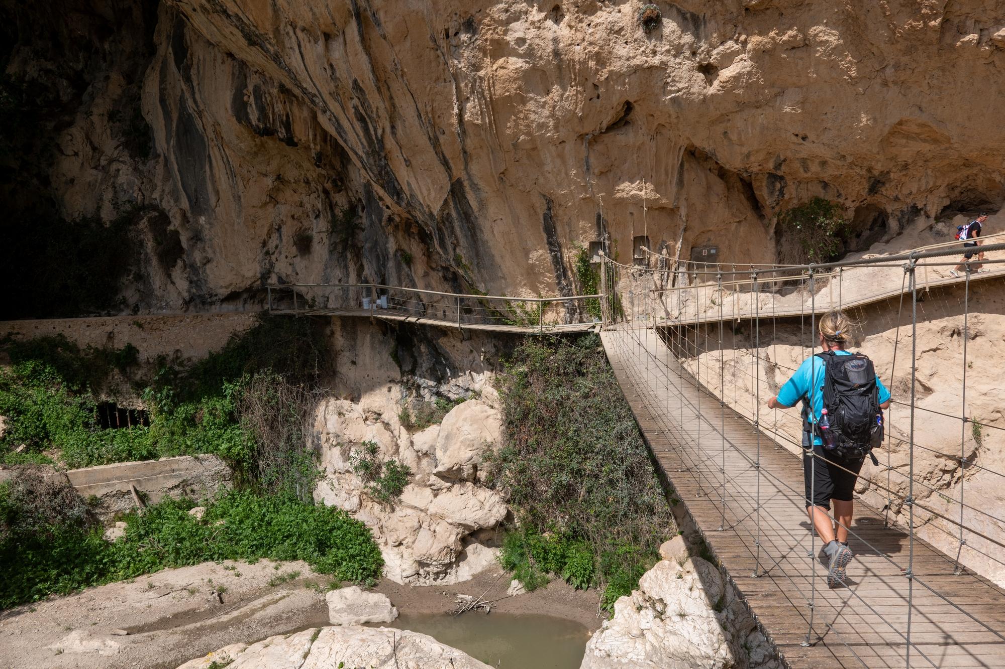 A person in blue crosses a wire/wood bridge above a river with huge cliffs above