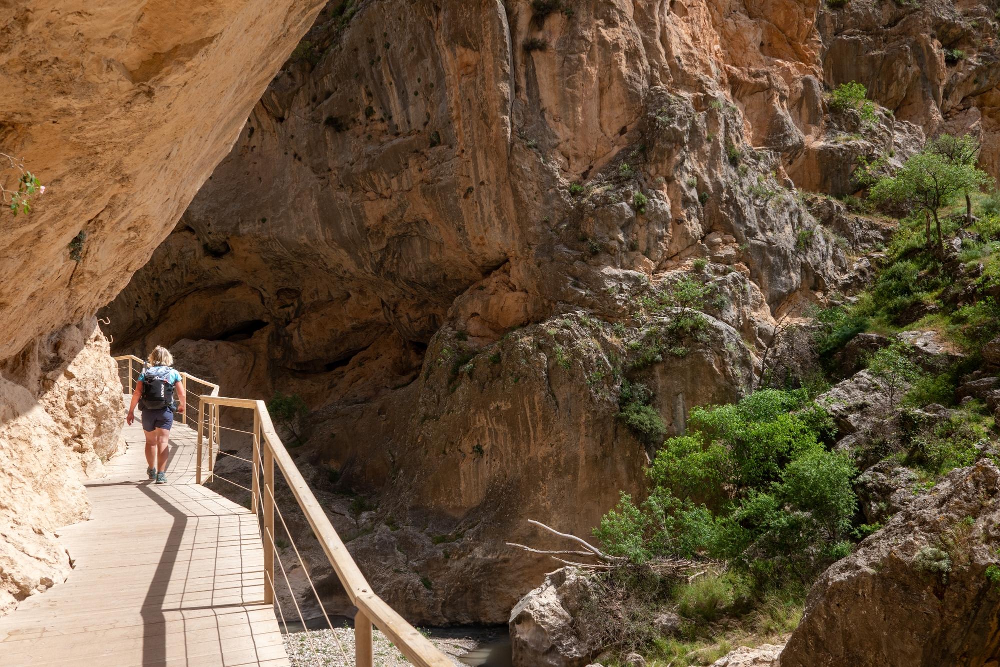 A hiker walks along a wooden elevated platform alongside cliffs and above a river