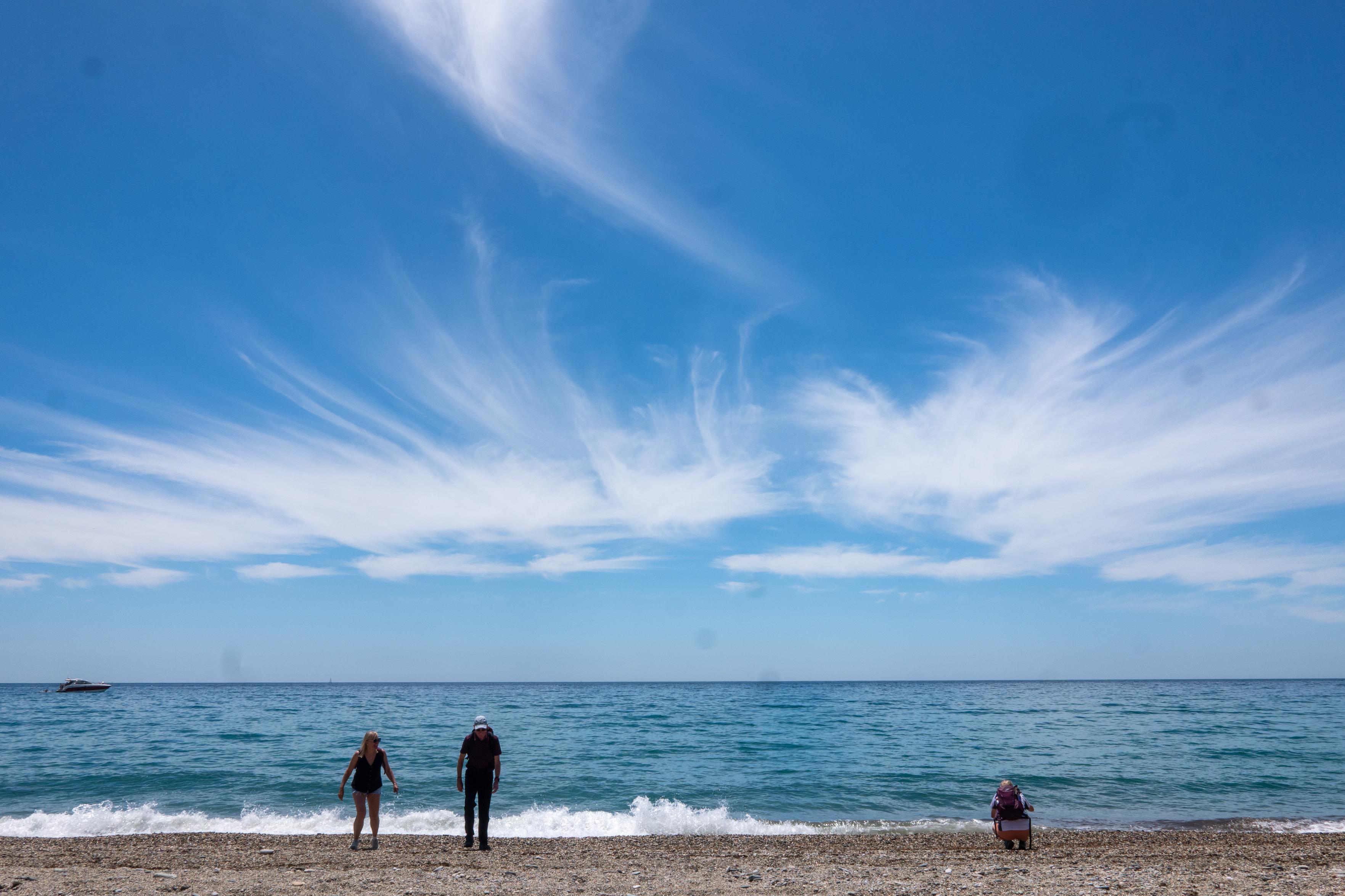 Three figures bottom of image watch the waves under a clear blue sky with whispy clouds