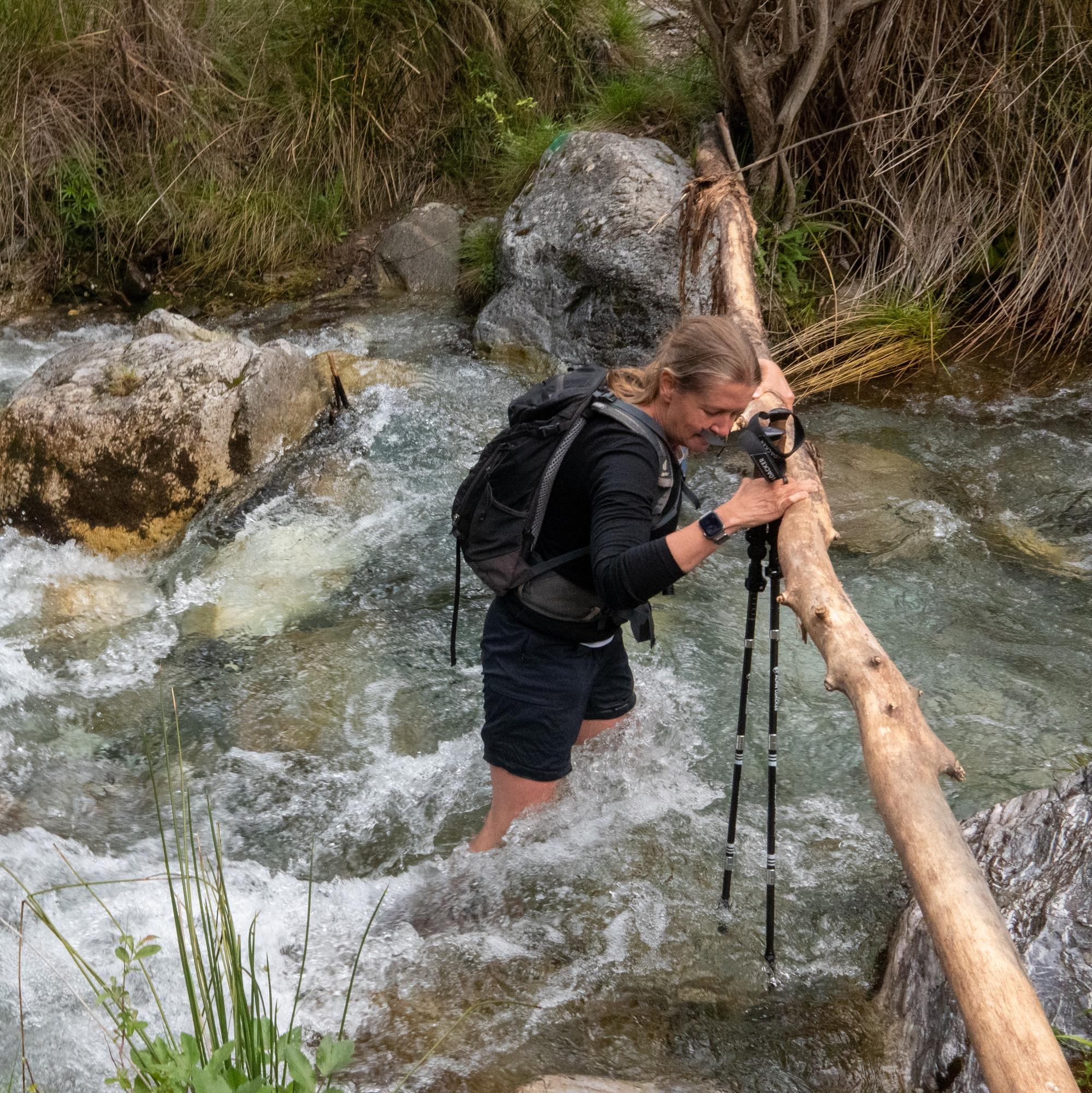 A hiker attempting to cross a fast flowing stream whilst holding onto a horizontal pole spanning the river