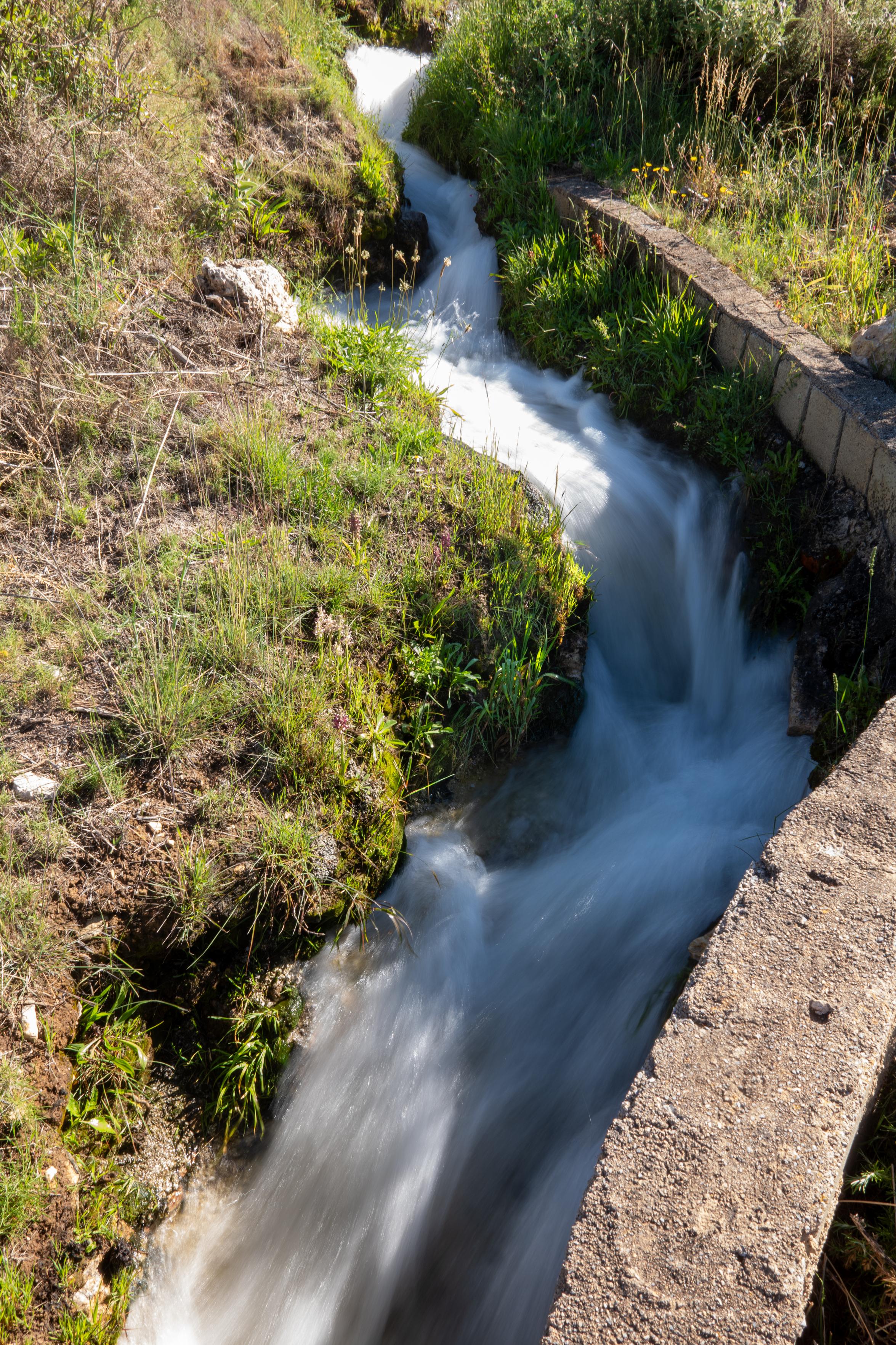 Water flowing fast down an irrigation channel. A wall to the right and green low grasses to the left