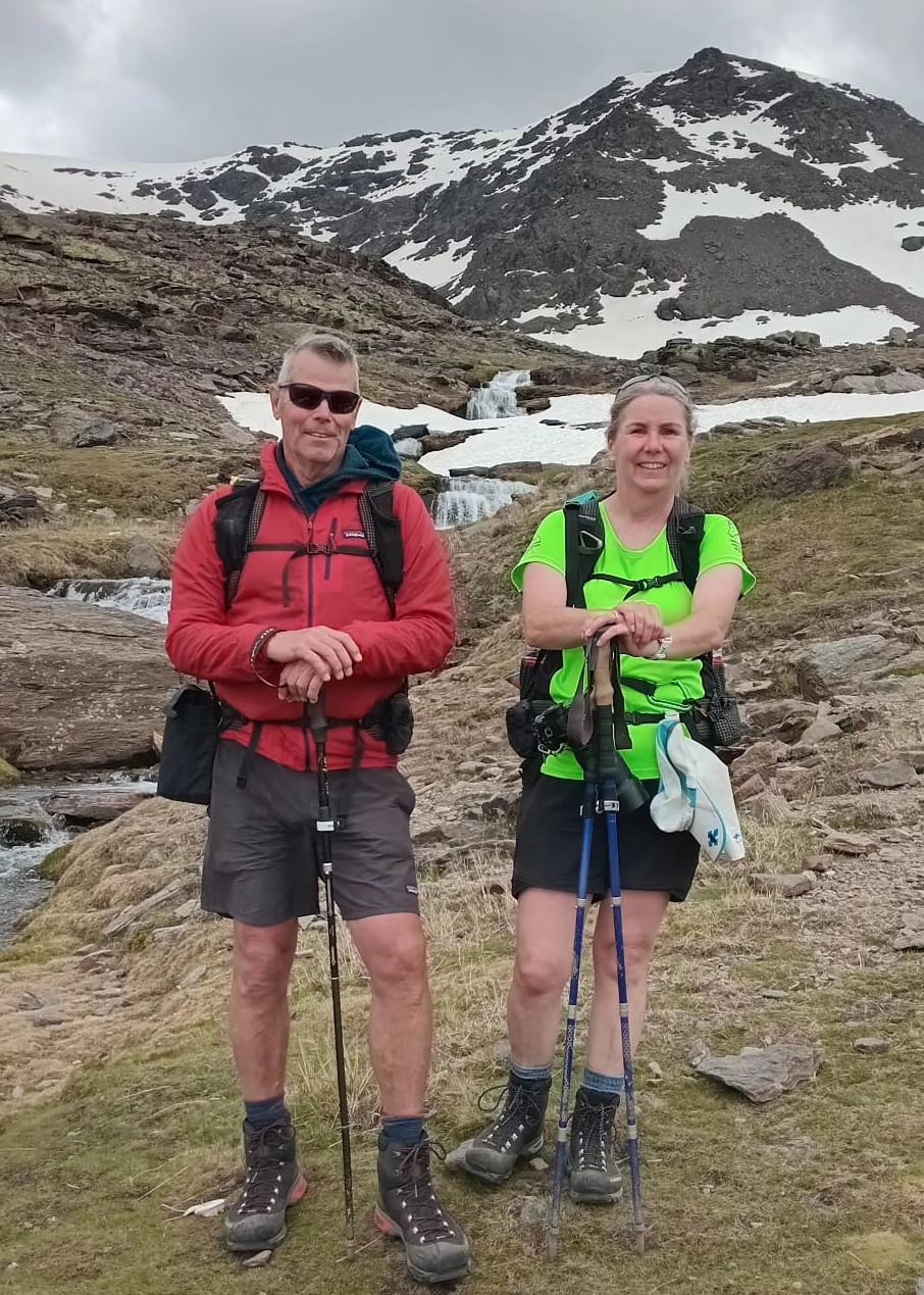 2 people in hiking gear pose for the camera with snow covered mountains behind