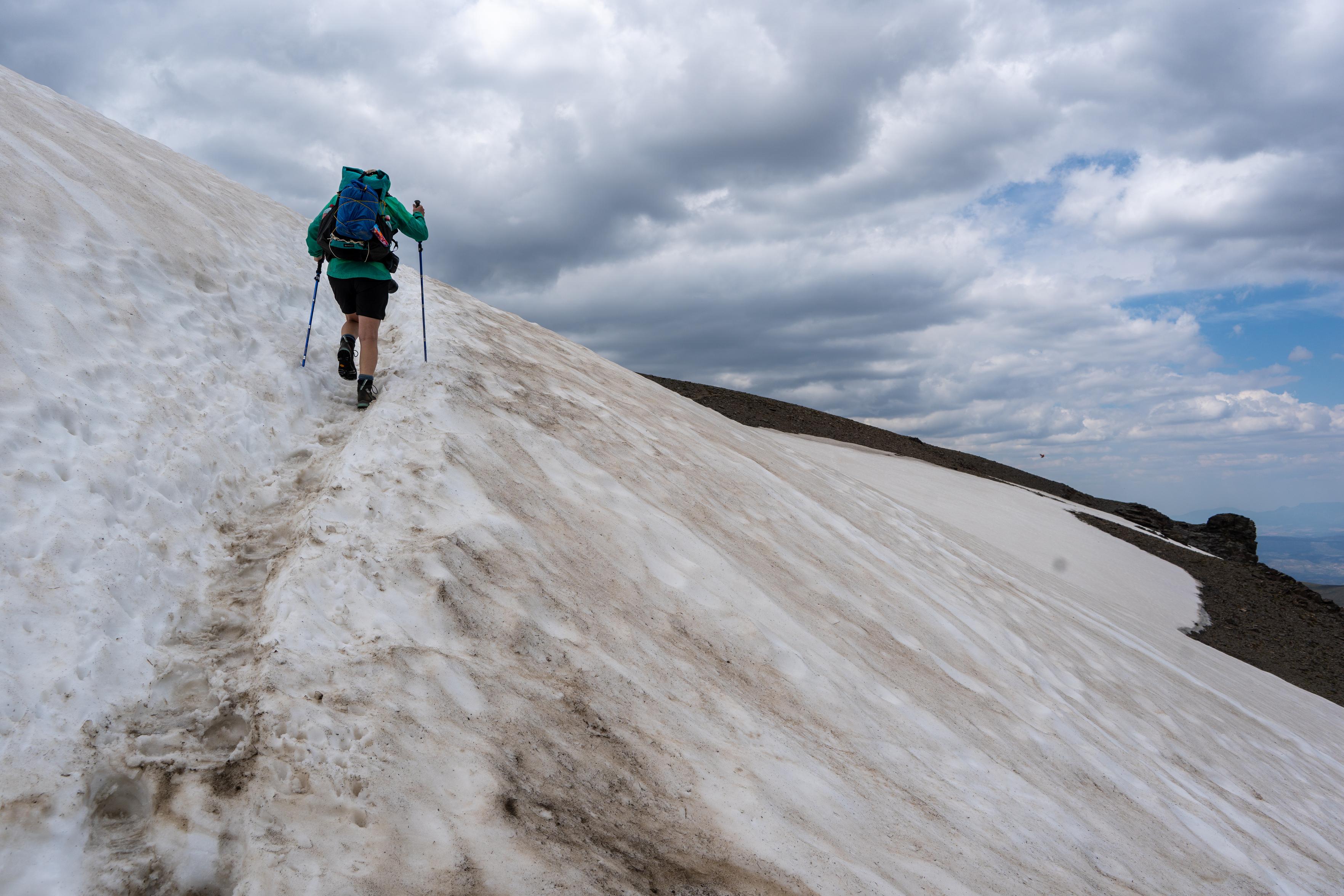 A person with green jacket in the top left ascends some snow slopes. The snow is quite dirty with grit