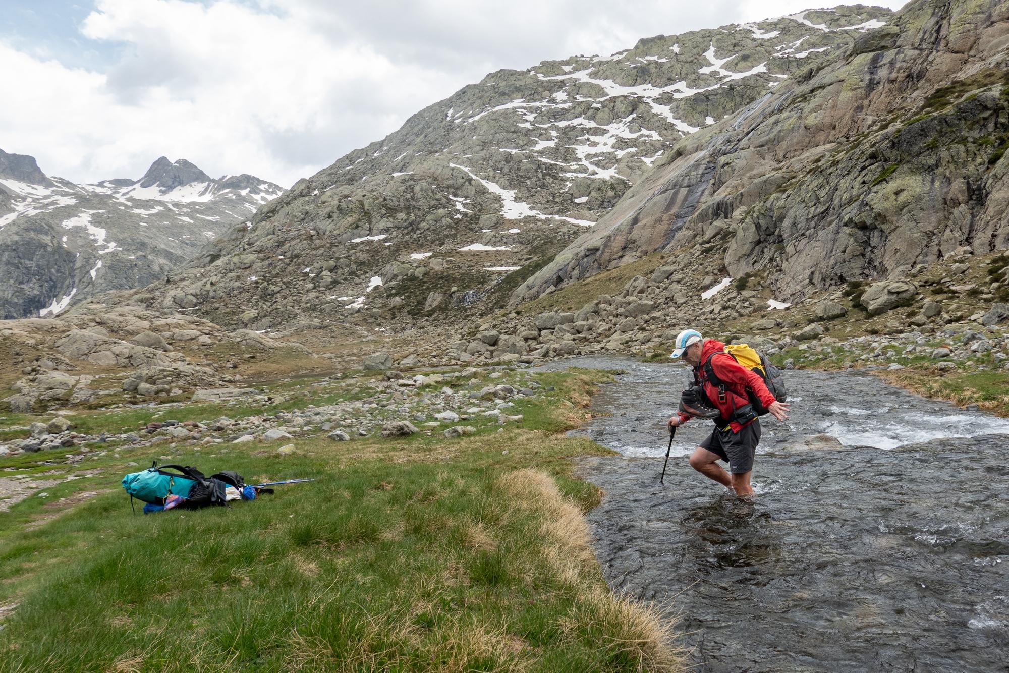 A person in red crossing a fast flowing river with boots strung around his neck