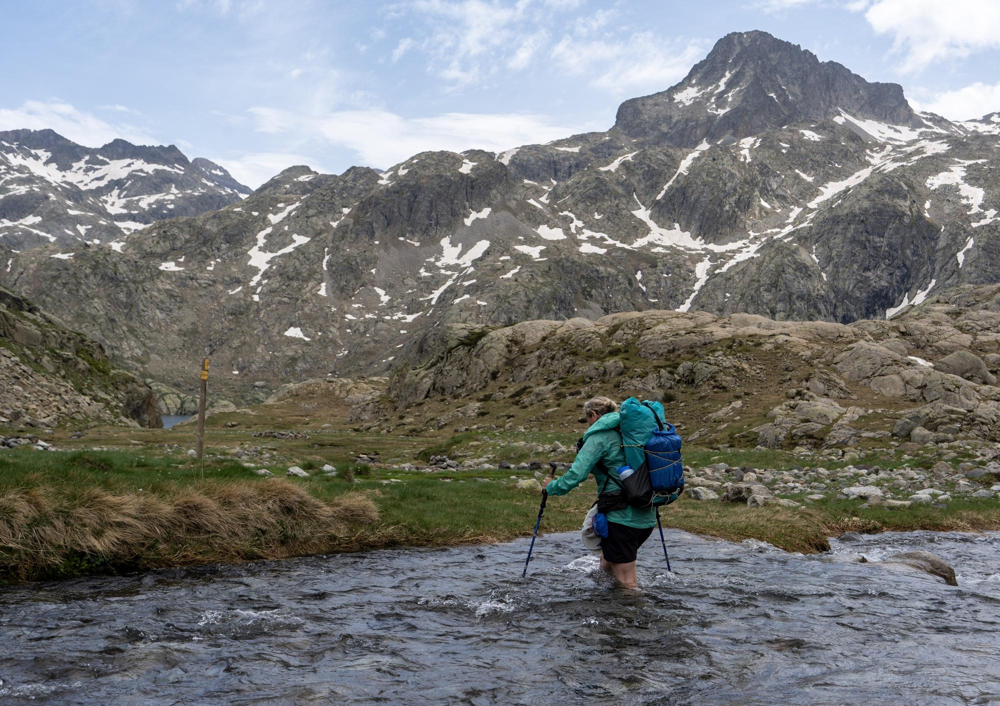 A person in a turquoise jacket and backpack is crossing a river