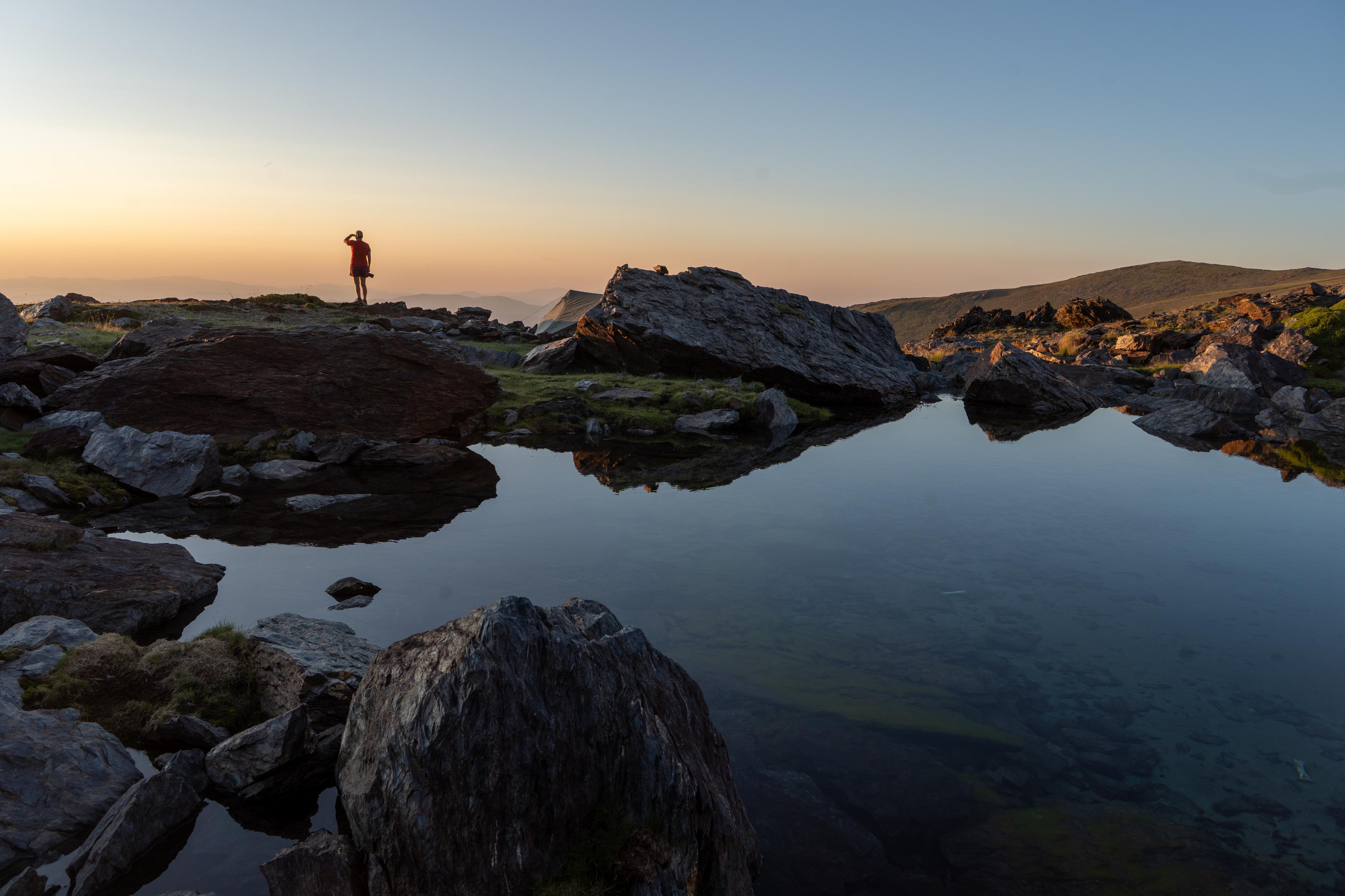 A hiker is illuminated by the sunset colors. In front is a still lake with large rocks and boulders