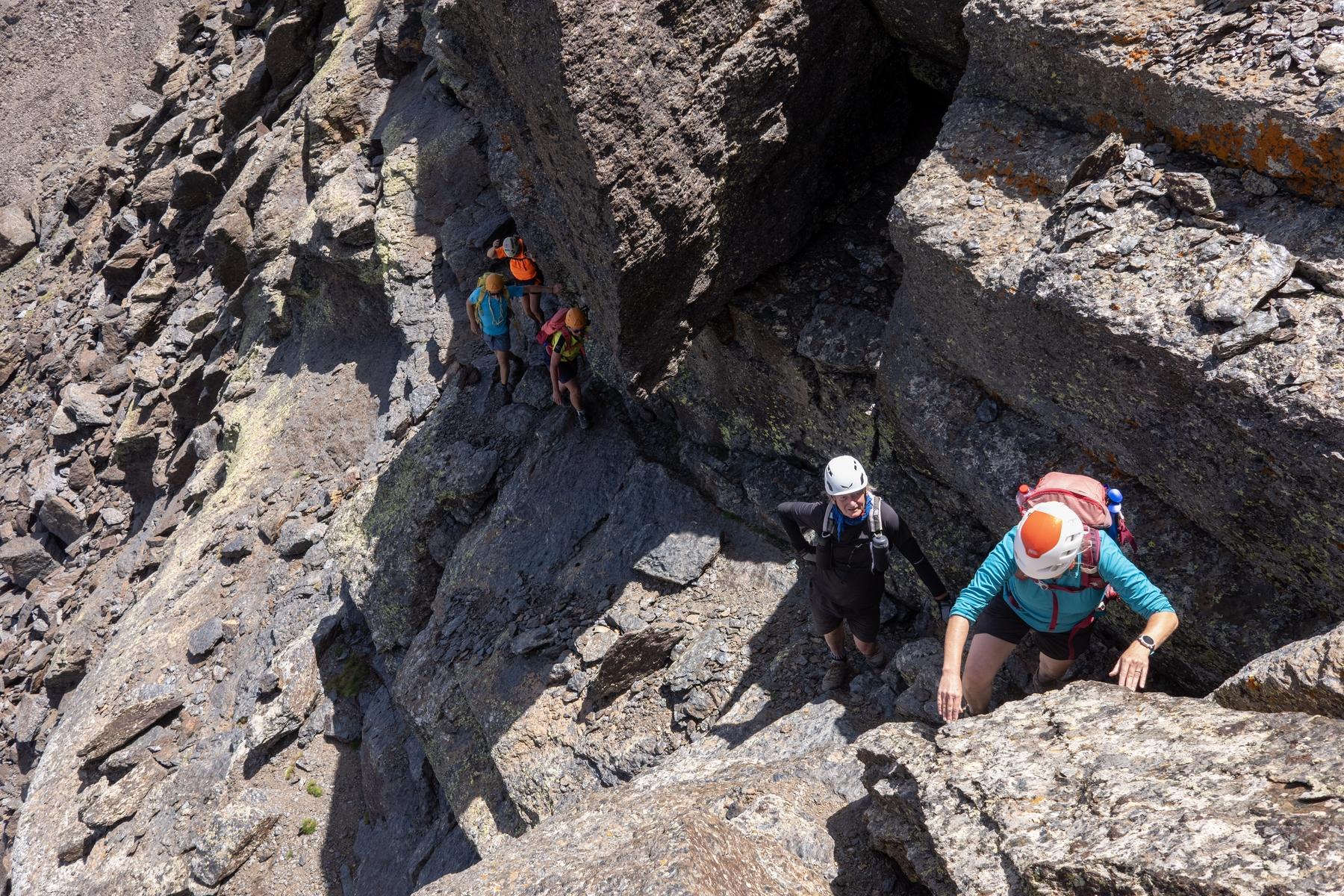 A group of people in colorful clothing and helmets pass along a narrow ledge with vertical rock above and below