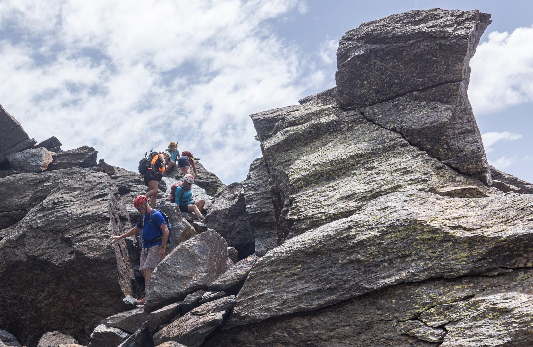 A group of helmeted hikers in colorful clothes clamber and scramble along a rocky and block strewn high level mountain ridge