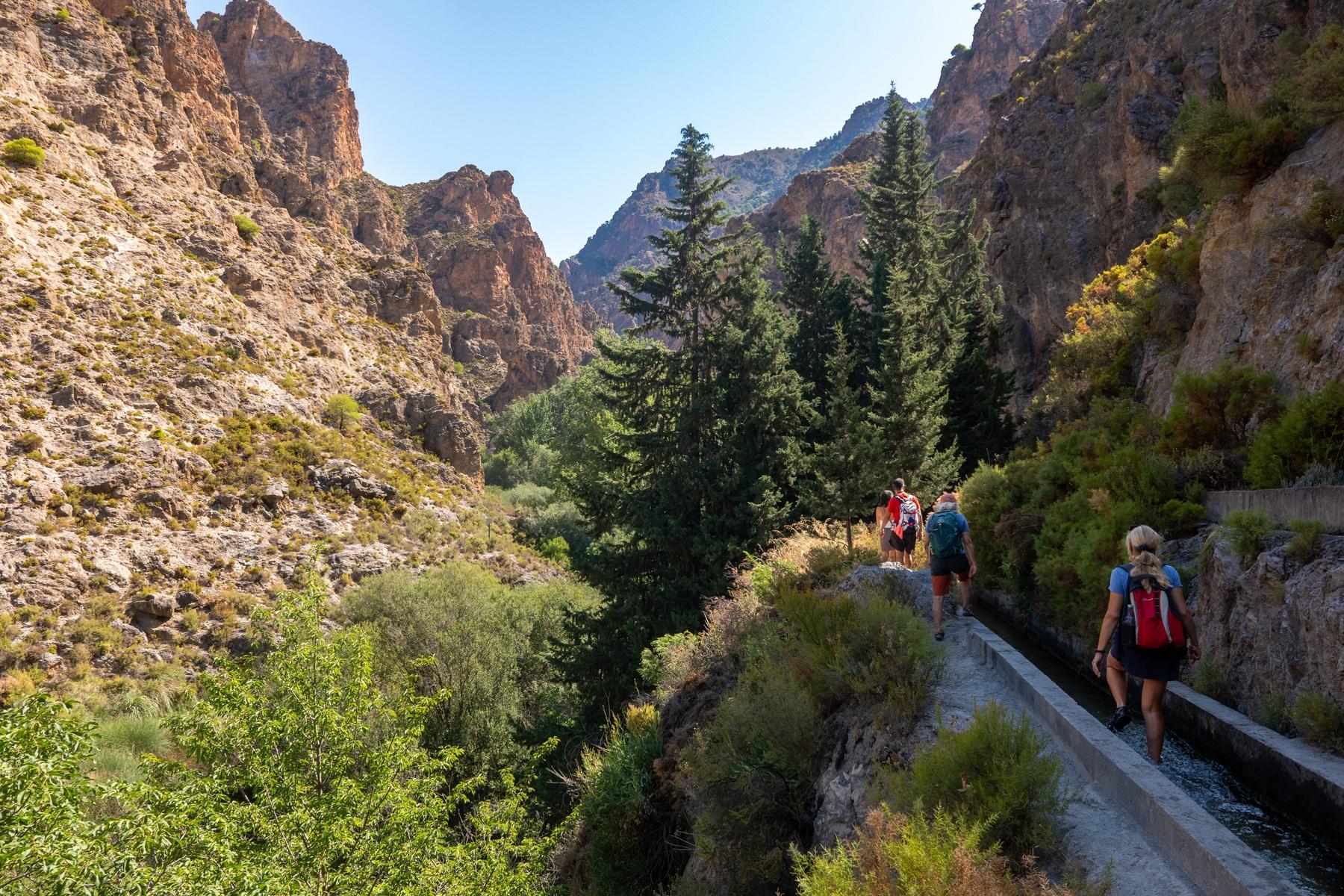 Some hikers walk along a narrow path on the right under a cliff. Ahead lies a gorge