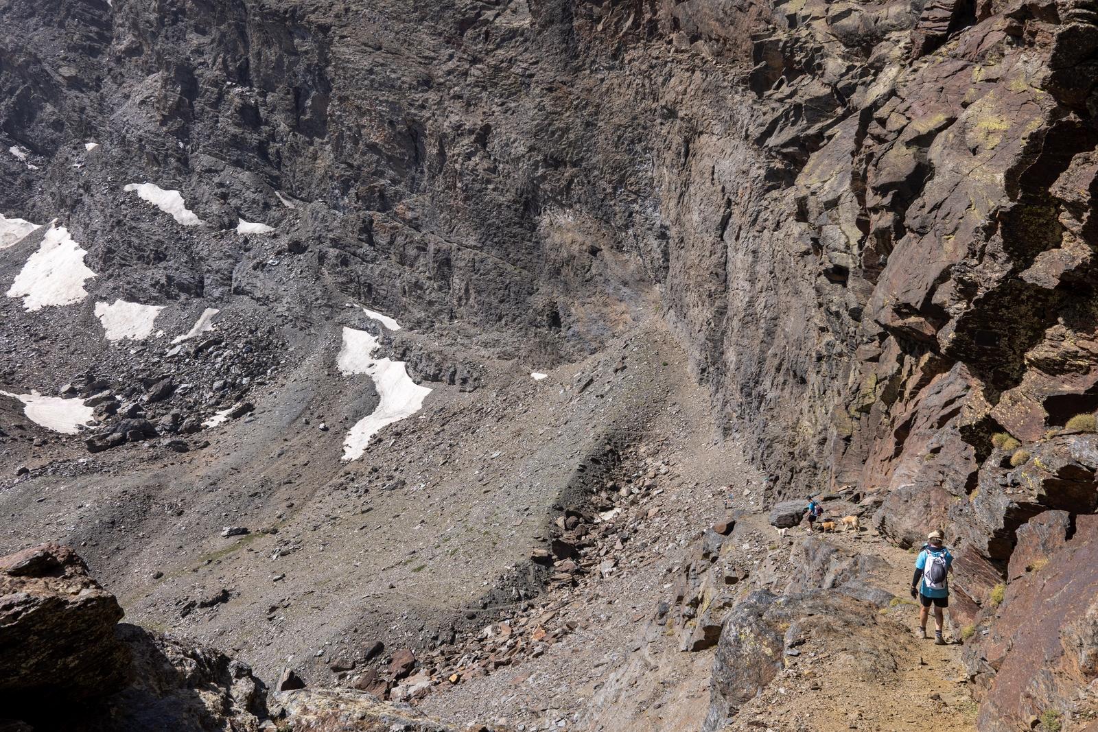 A person in light blue descends a track on the right with huge rock walls towering above. There are still snow patches lower down in the valley
