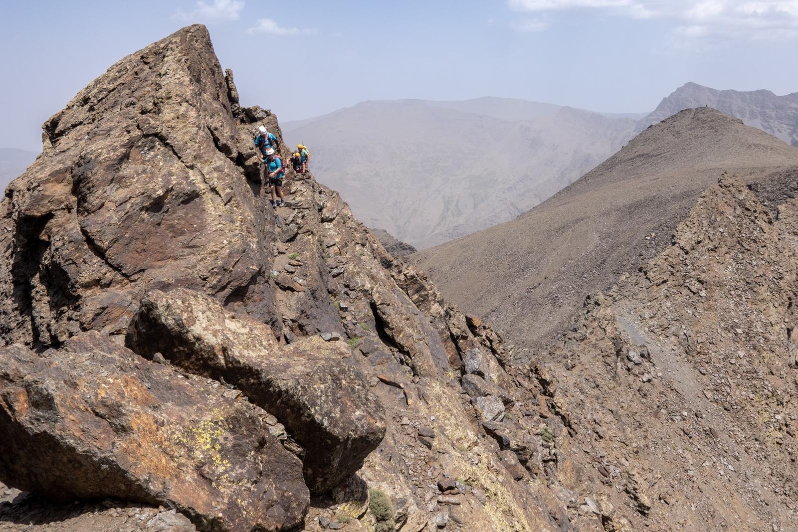 Some people cling to a narrow ledge on the side of the peak of Salon. Mountains stretch away to the right