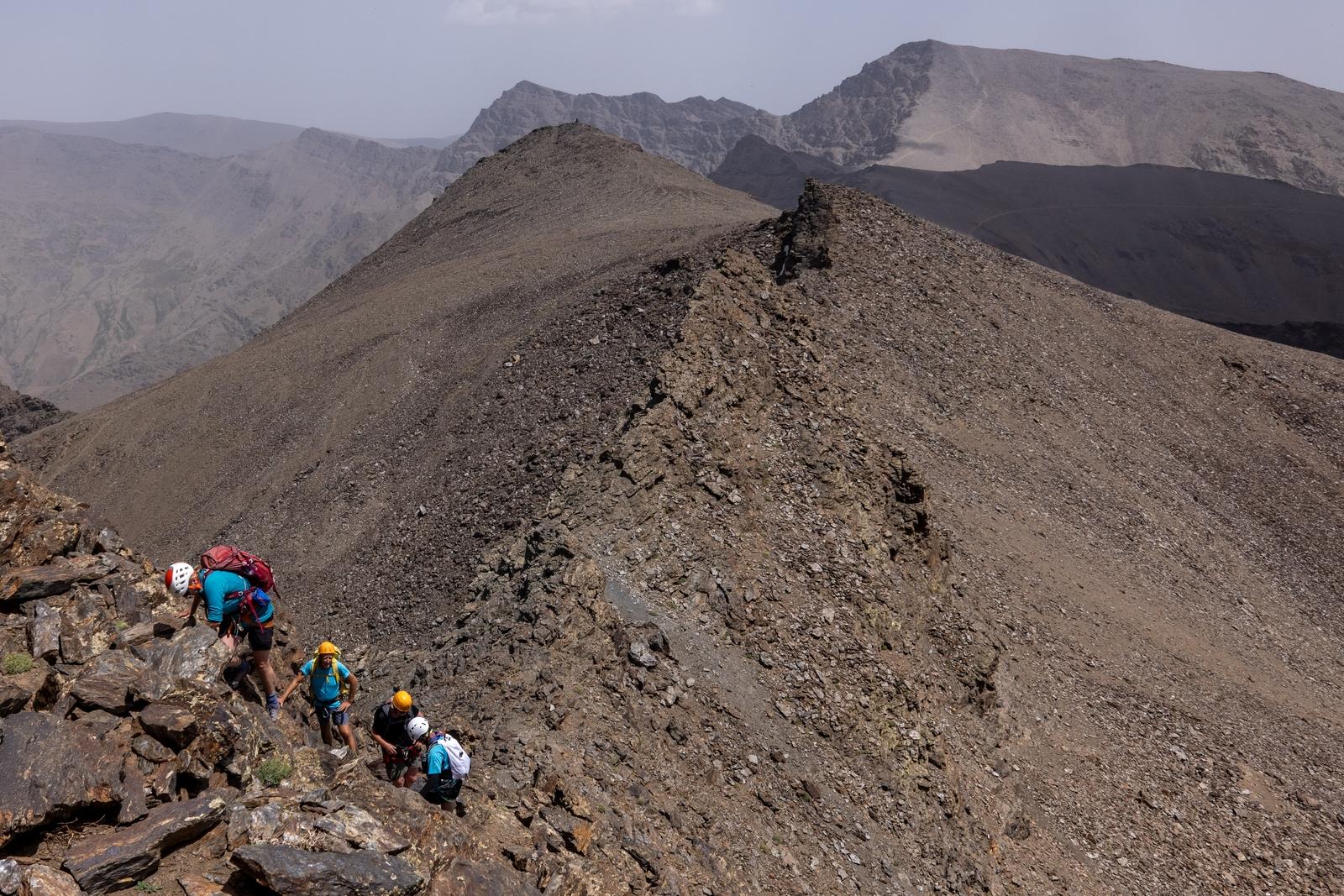 4 scramblers in brightly colored gear and helmets stand out on a brown rock mountain face with more mountains (Alcazaba and Mulhacen) stretching away into the background to the right