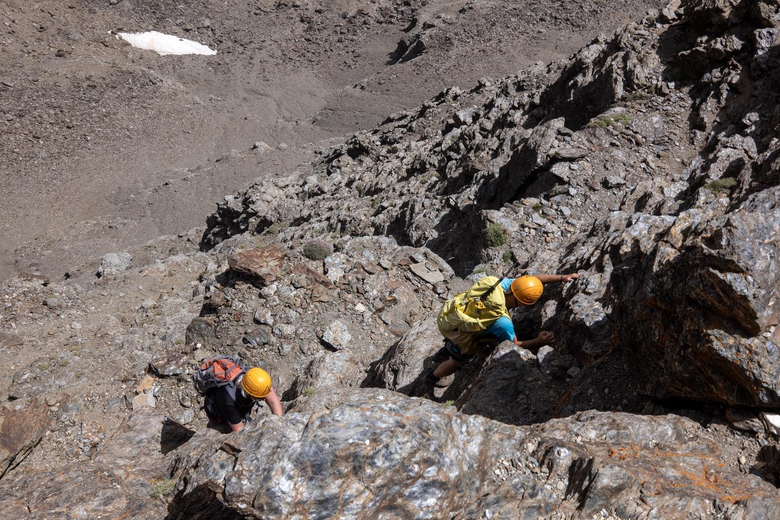 2 scramblers in brightly colored gear and orange helmets start climbing a short but steep rock wall