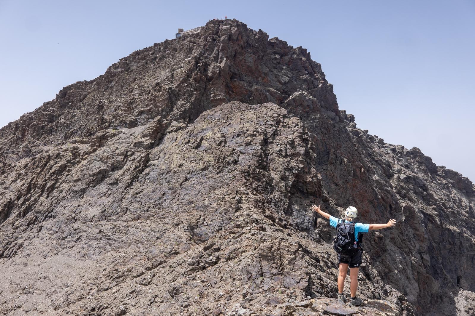 A person in blue with white helmet stands below the summit of Veleta with arms outstretched