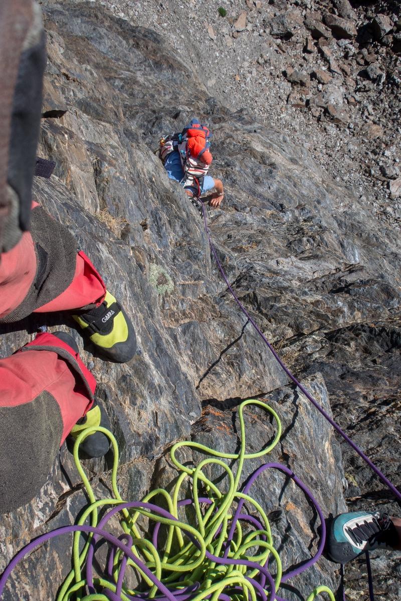 Two people stand on small footholds, a yellow and purple rope between them on the floor. vertically below is a rock climber with light blue t-shirt climbing up