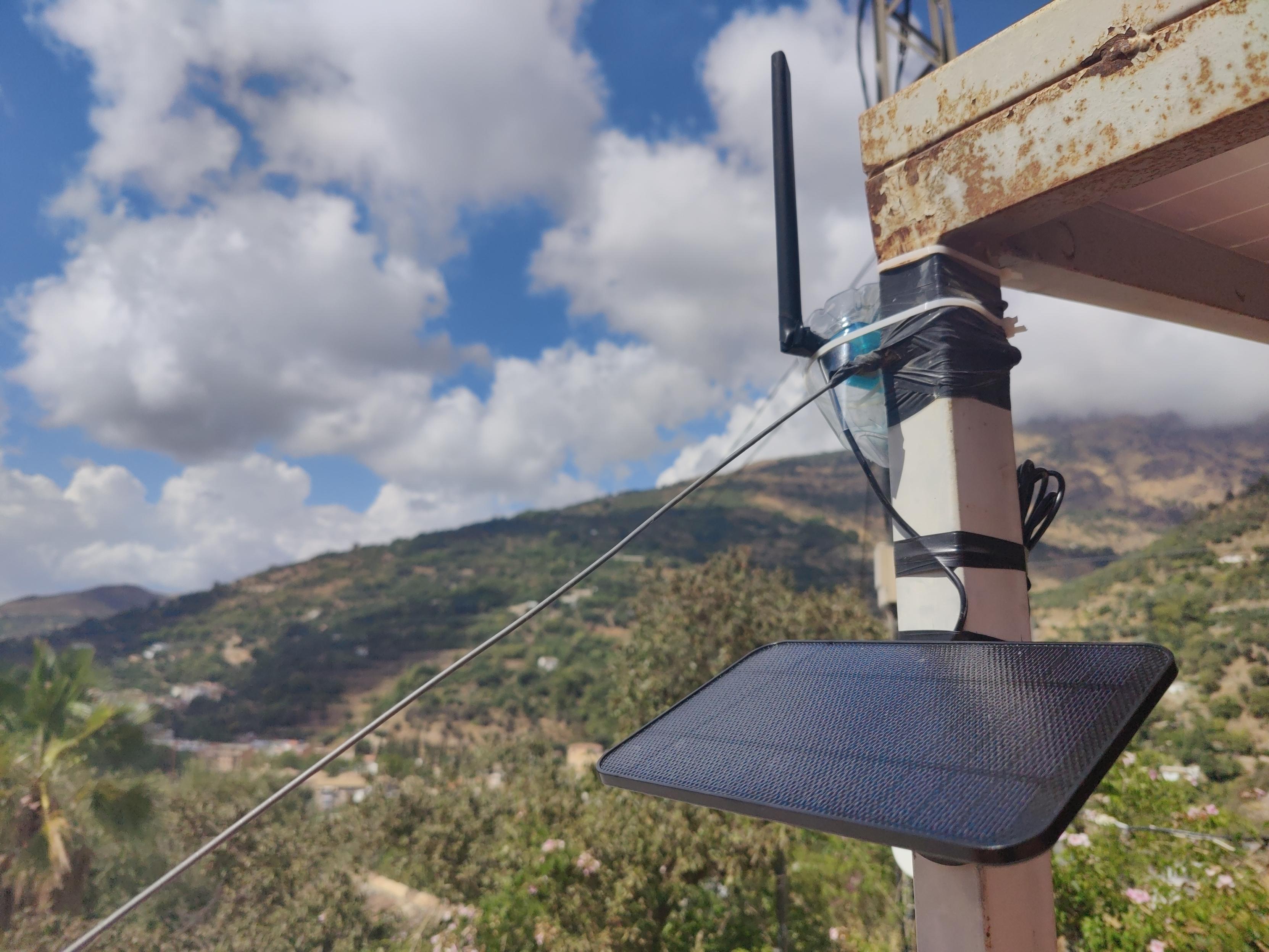 A solar panel sits suspended below an old water bottle containing a small radio. A black antenna protrudes from the bottle. Green hillsides beyond. White clouds in a blue sky