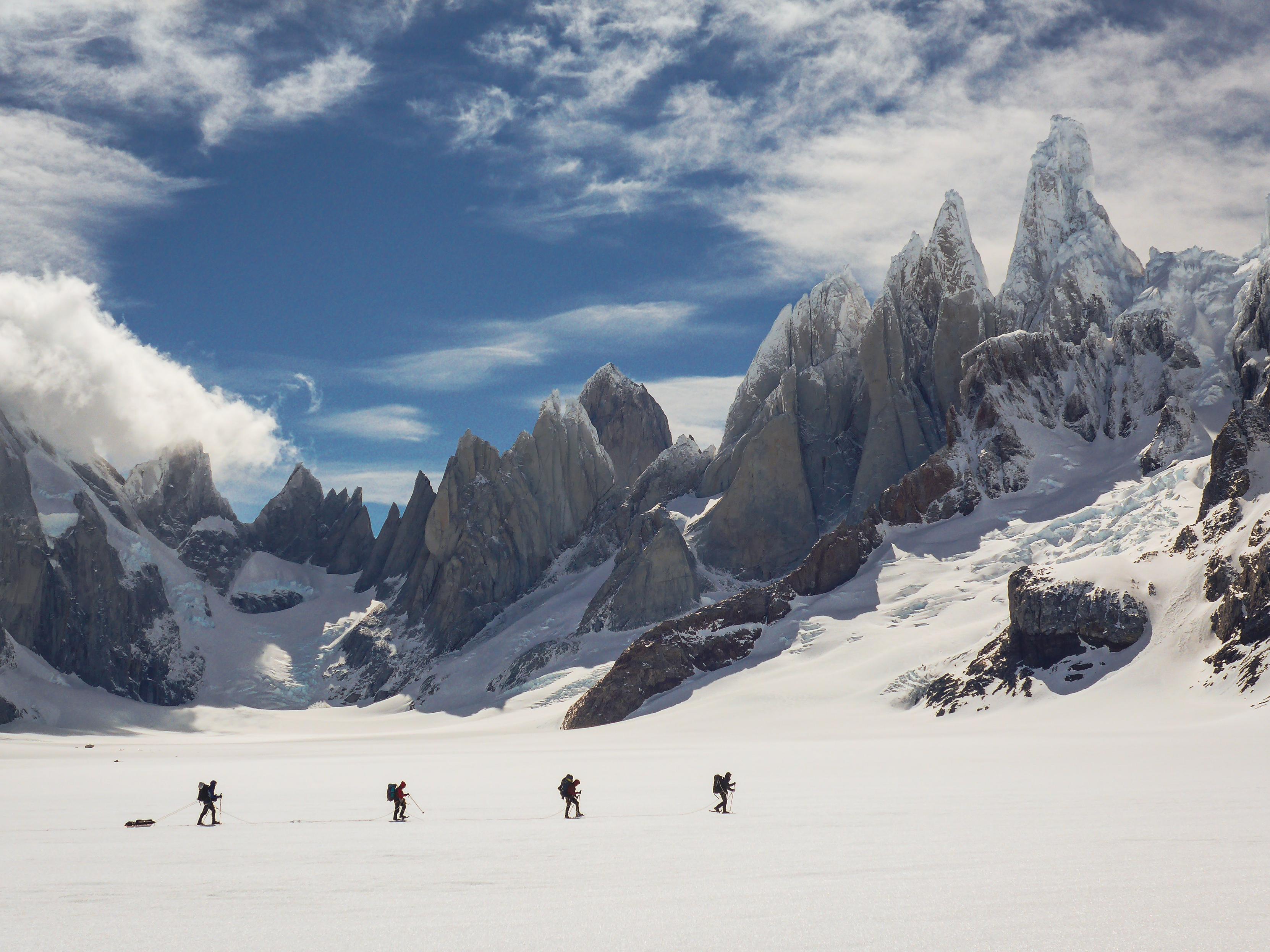 4 people, one pulling a sledge cross in front of a jagged mountain range of rock and ice. A gap in the white clouds reveals blue sky.