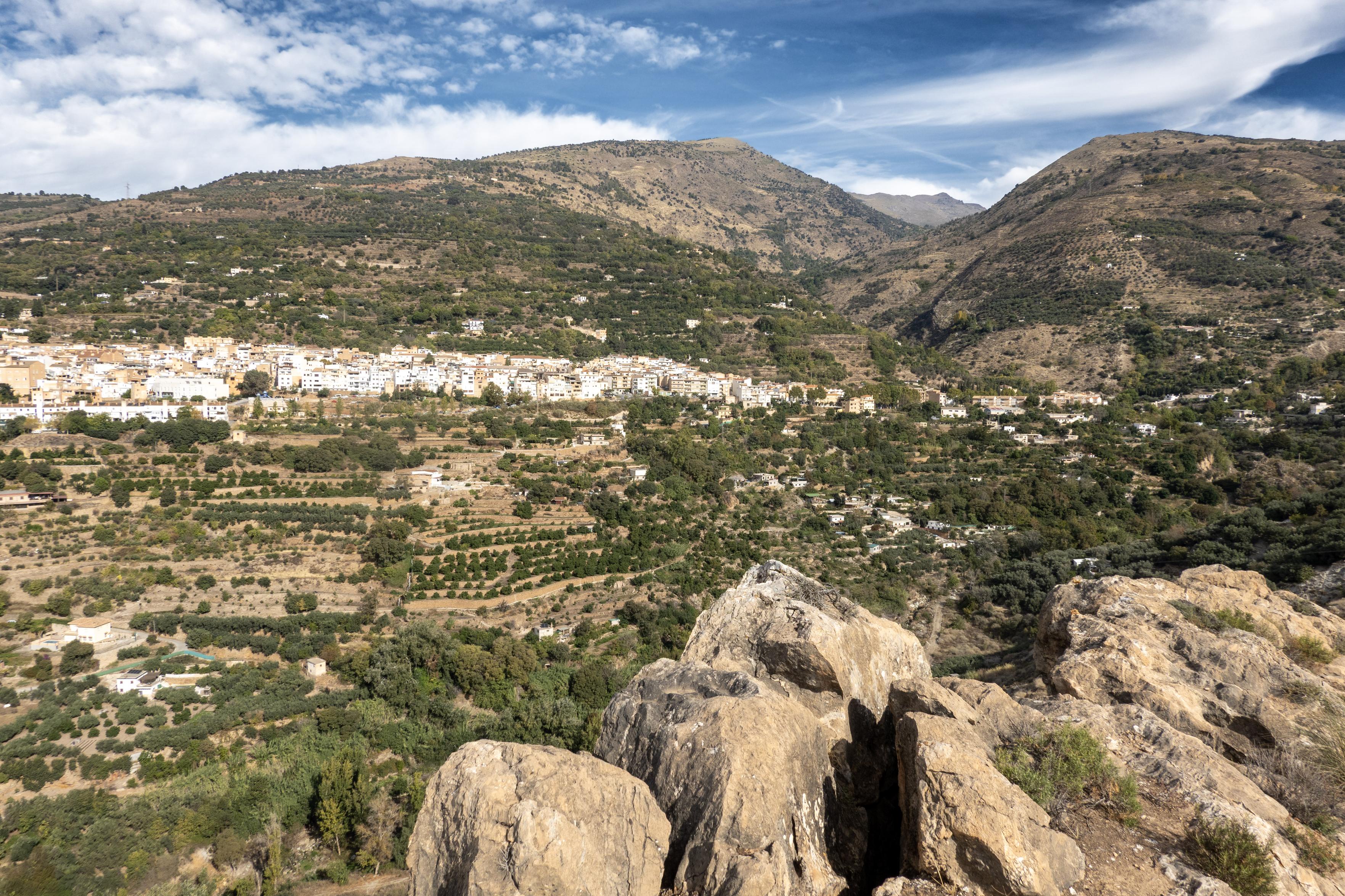 Sandstone rocks in the foreground. Behind a white village passes horizontally across the hillside. behind rise higher mountains with a deep river valley to the right