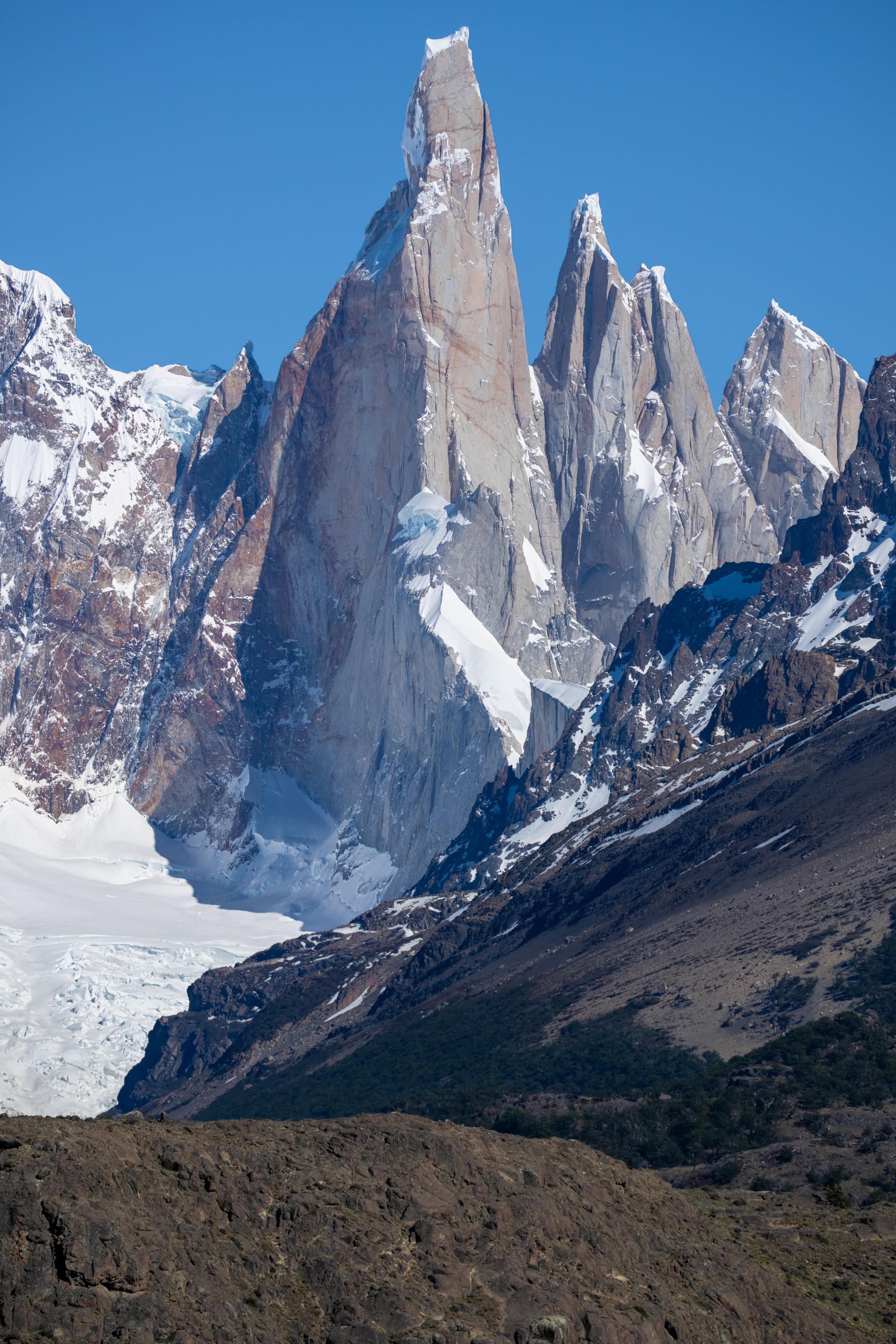 A sharp needle of a mountain rises vertically into blue sky. Formed of granite the rock has orange tint. Glaciers at its foot