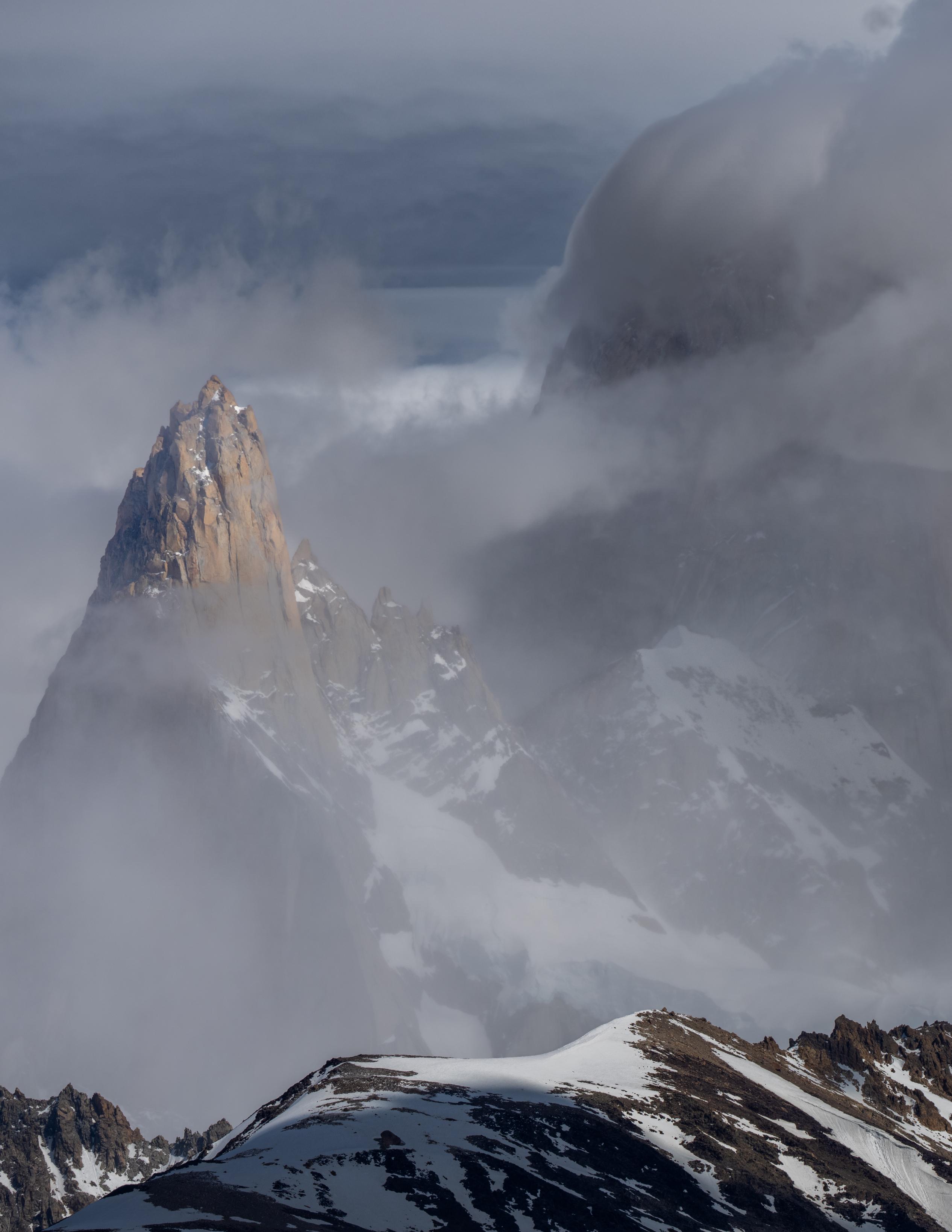 The orange granite rocks of Aguja Poincenot summit appear out of thin cloud. To the right Fitzroy looms dark and menacing. The foreground has a level ridge of rock and snow